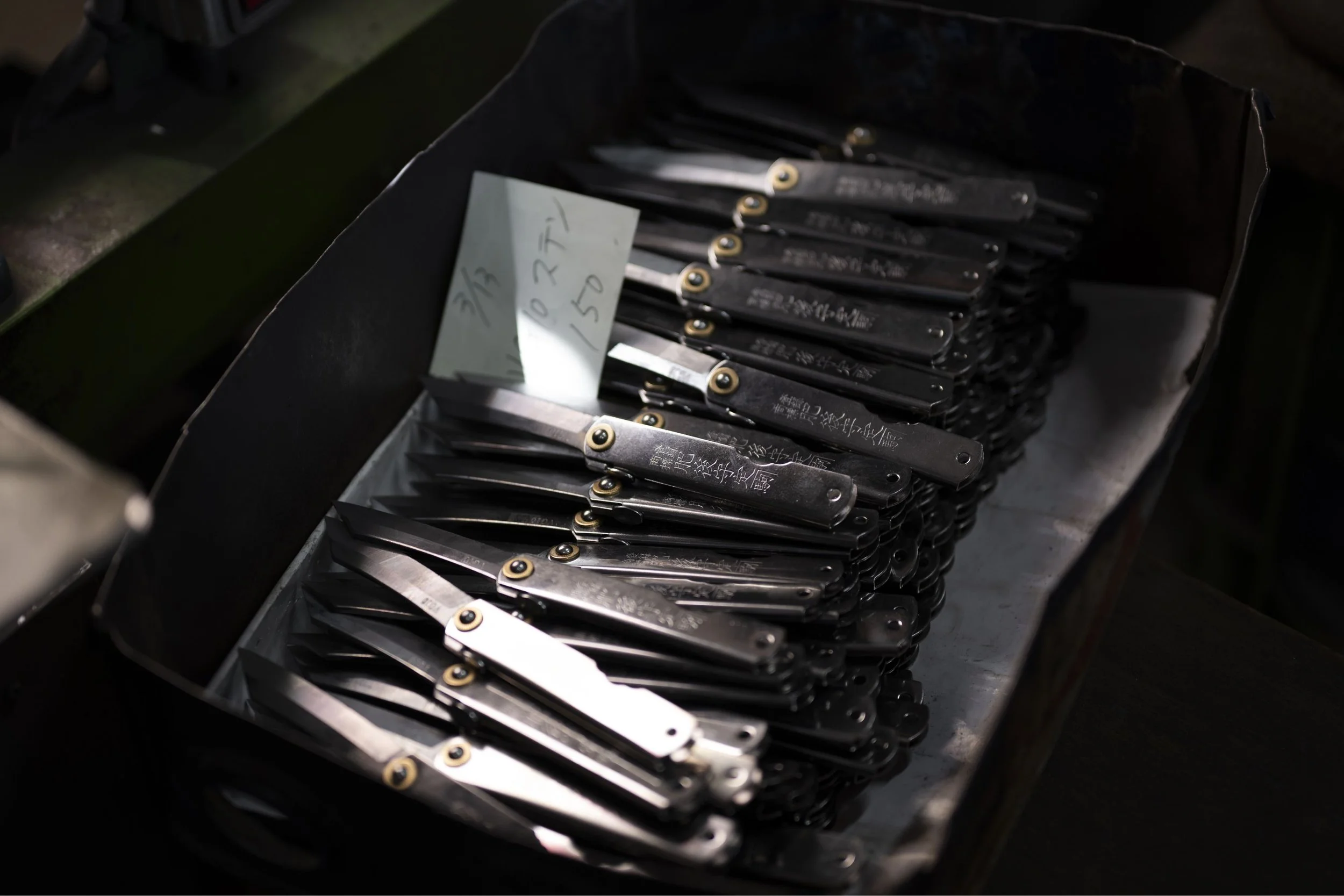 A box of black folding knives with gold accents and Japanese inscriptions, with a handwritten note in Japanese on a sticky note inside the box.