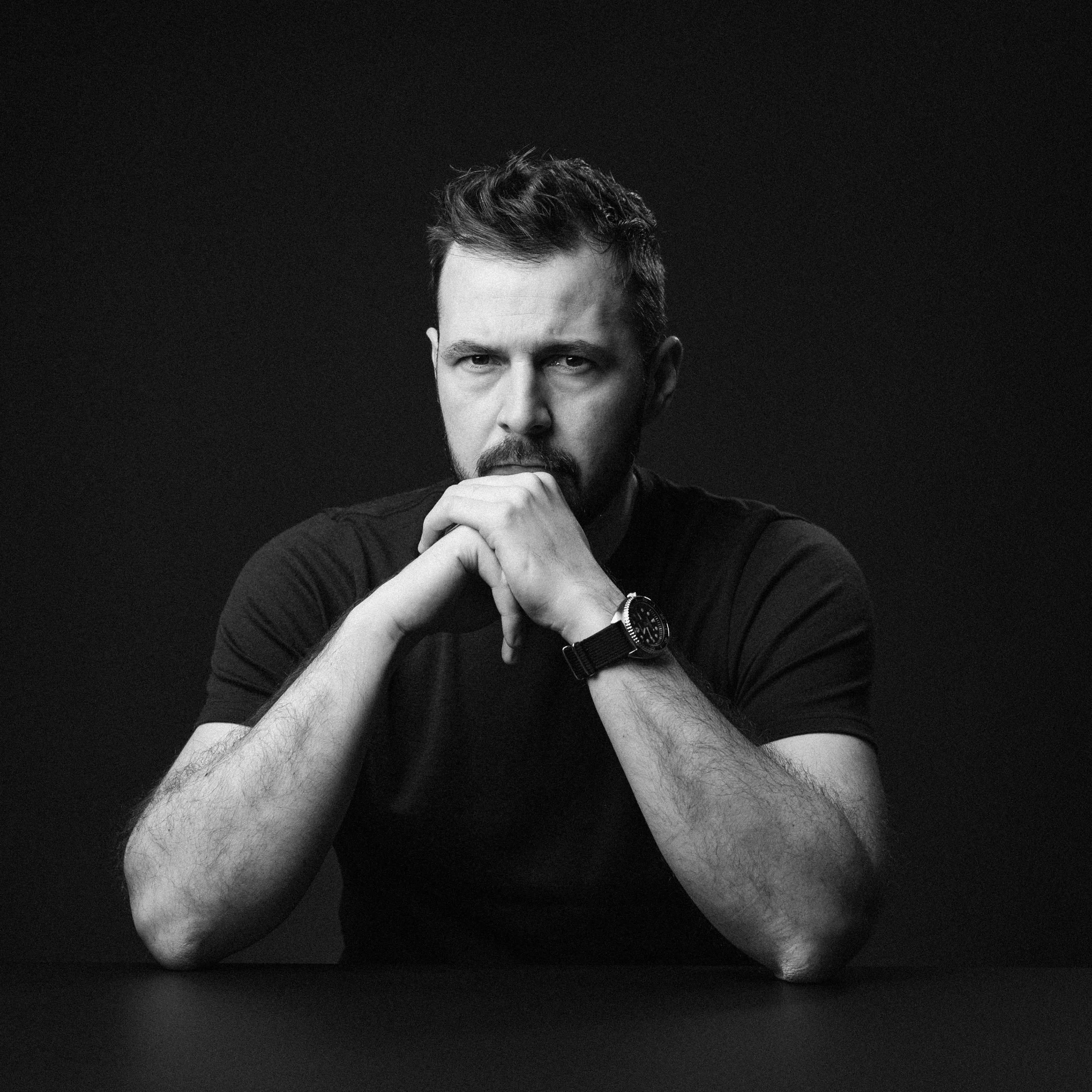Black and white portrait of a man with short hair and facial hair, resting his chin on his left hand, wearing a watch, against a dark background.