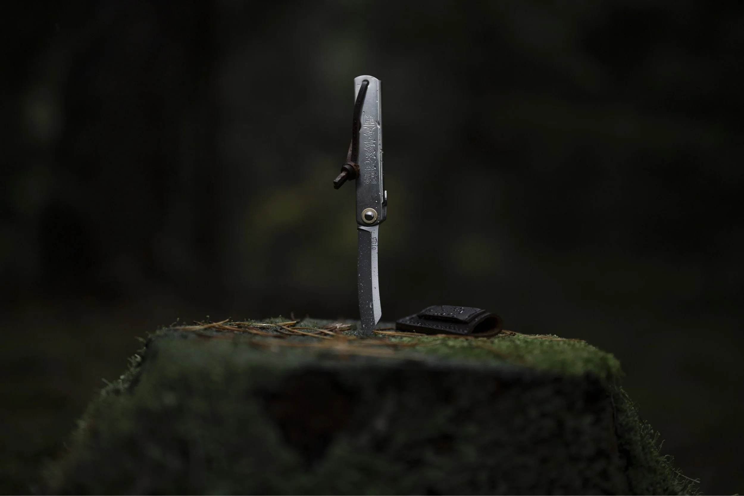 A pocket knife standing upright on a moss-covered surface with a dark background.