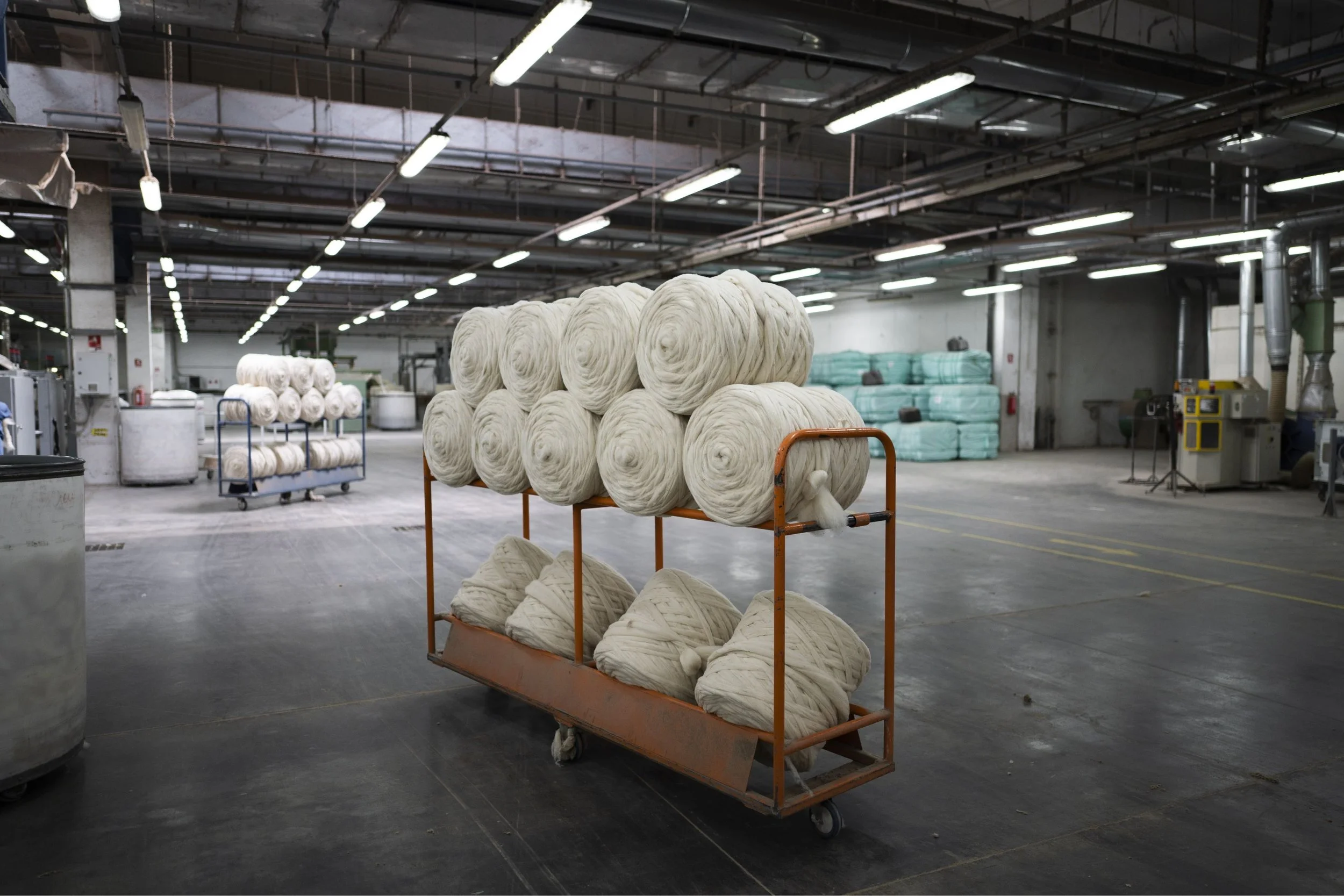 A warehouse holding large spools of white yarn on an orange wheeled cart. The warehouse has industrial lighting, exposed ductwork, and additional large bundles of yarn stacked along the walls.