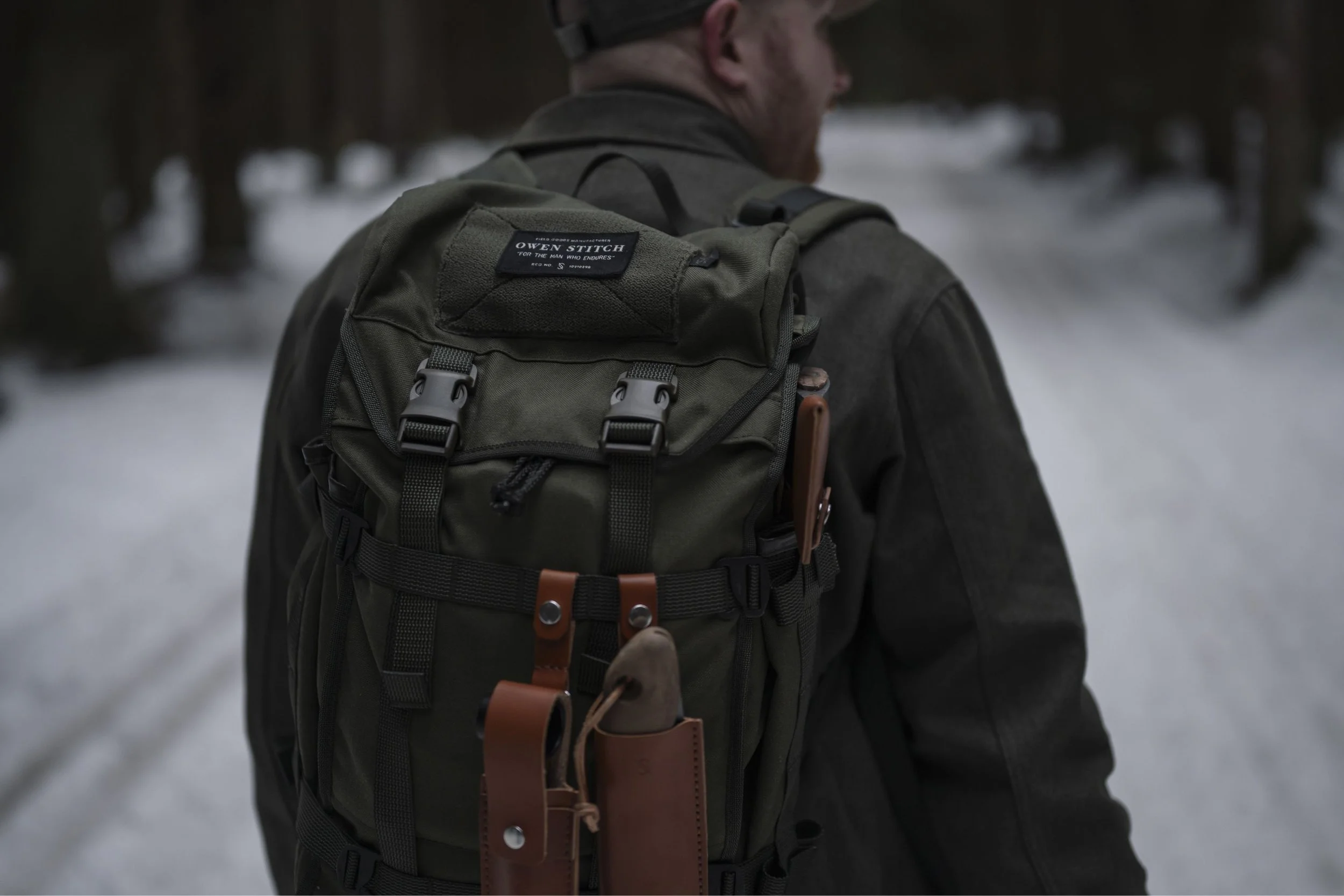 A person walking outdoors in winter, wearing a dark jacket and carrying a large olive green backpack with black and brown straps.