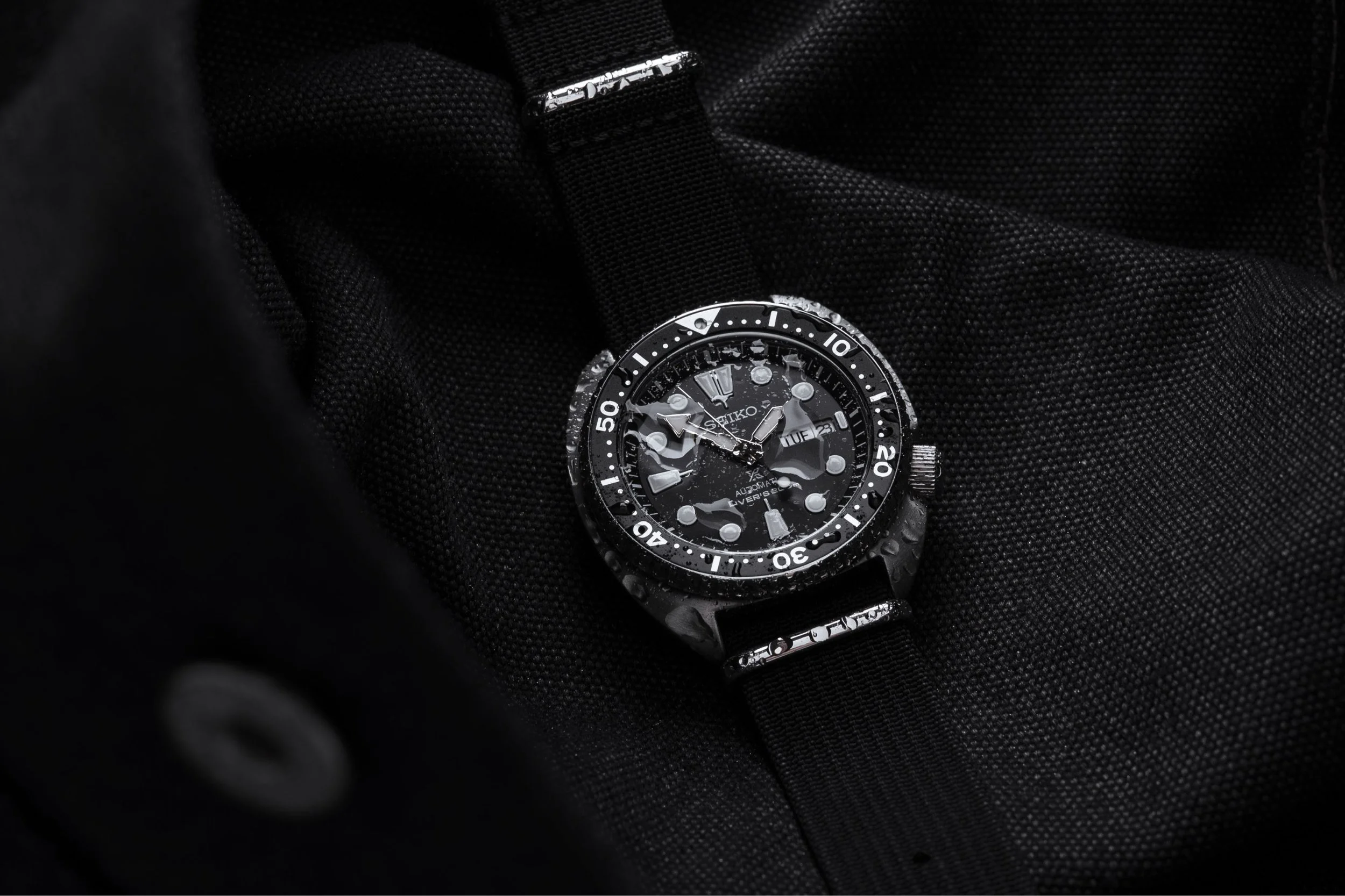 A black Seiko dive watch with a black strap resting on a black textured fabric surface, with water droplets on the watch face and strap.