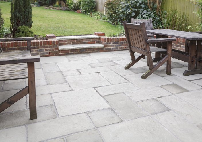 Backyard patio with stone pavers, wooden outdoor furniture, brick stairs leading to a grassy yard, surrounded by greenery and bushes.