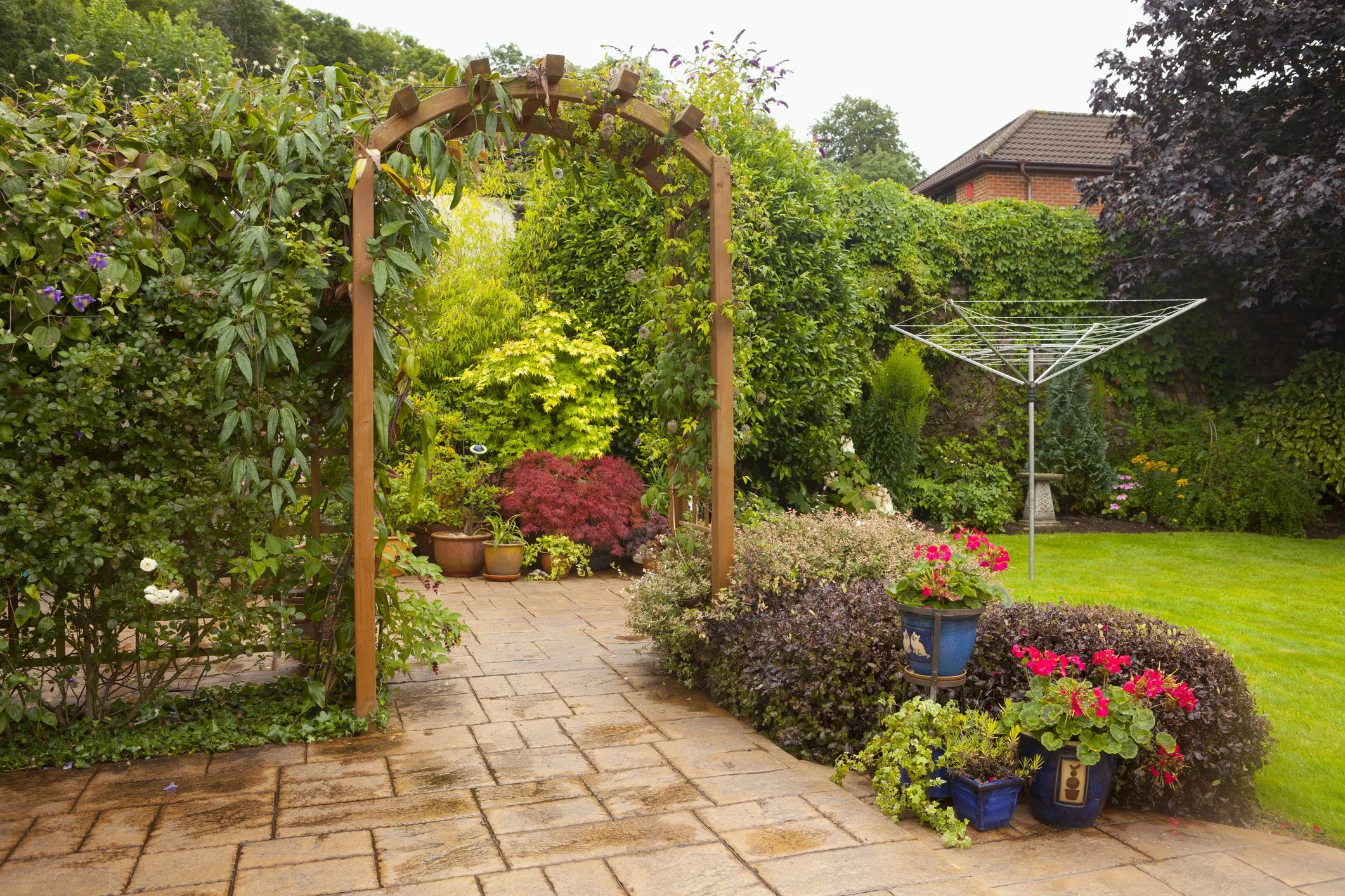 A lush garden with a wooden archway, potted plants, flowering bushes, a winding cobblestone path, a rotary clothesline, and a red brick house in the background.