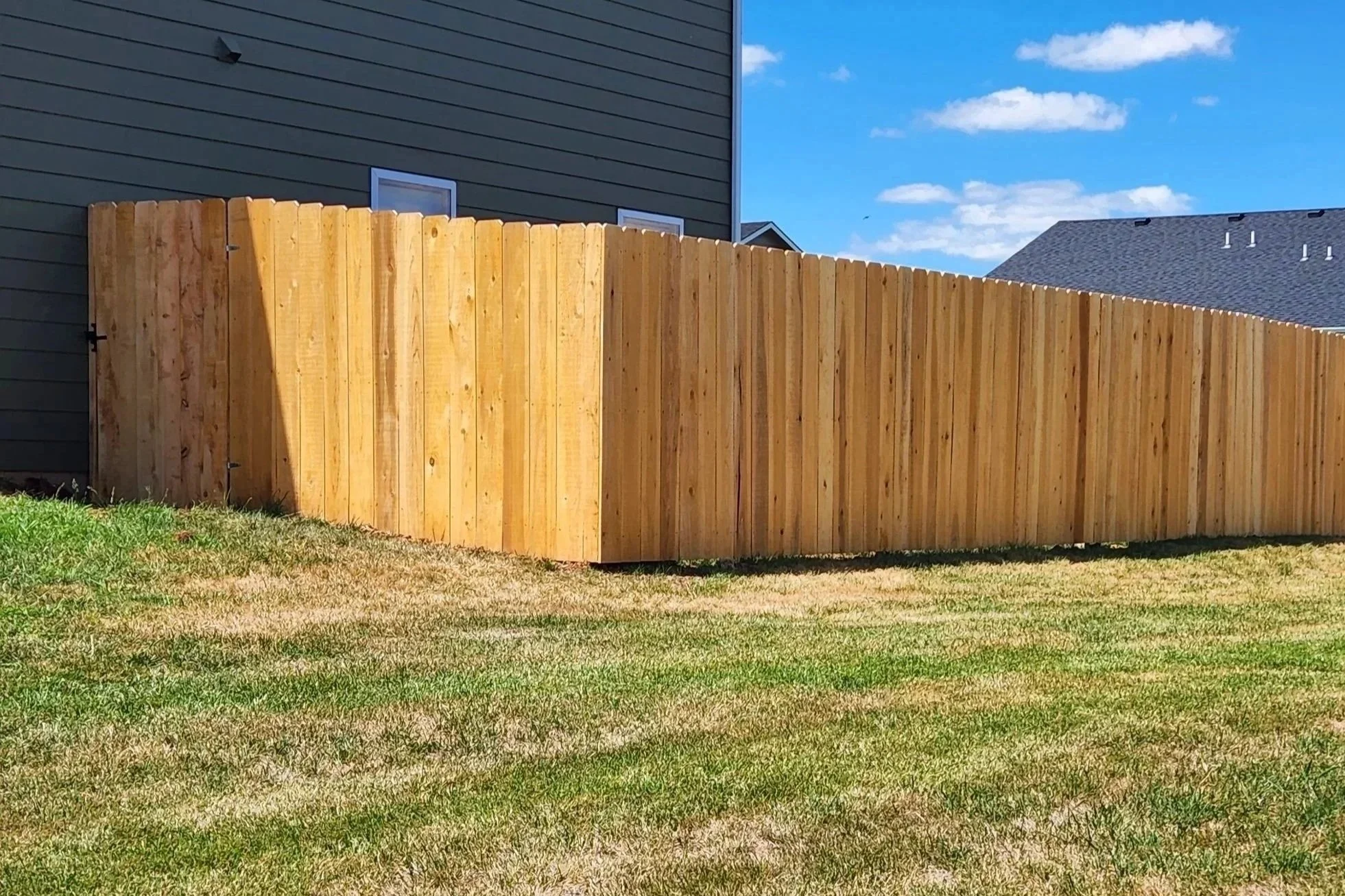 A wooden fence surrounds a backyard with green grass. In the background, there are two houses, one with dark grey siding and the other with a lighter roof. The sky is blue with some white clouds.
