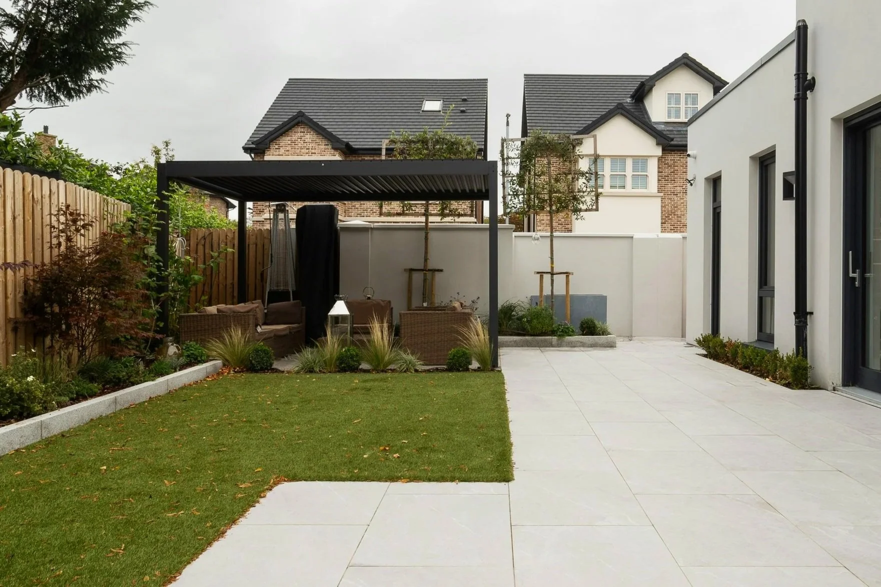 A modern backyard with a small lawn, a paved patio, garden beds with plants, a seating area under a black pergola, and neighboring brick houses with pitched roofs.