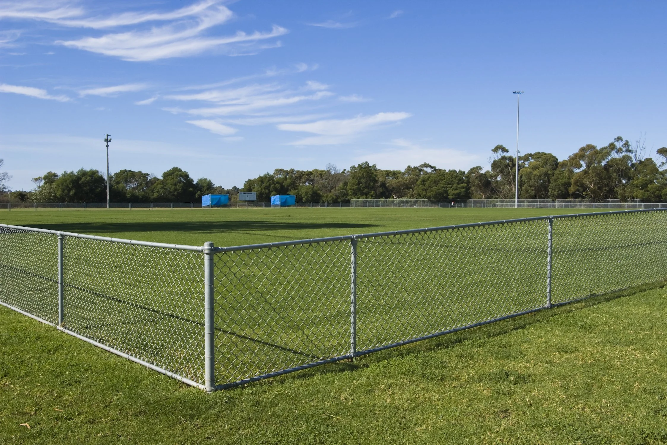 A sports field surrounded by a chain-link fence with a grassy area and trees in the background under a blue sky with scattered clouds.