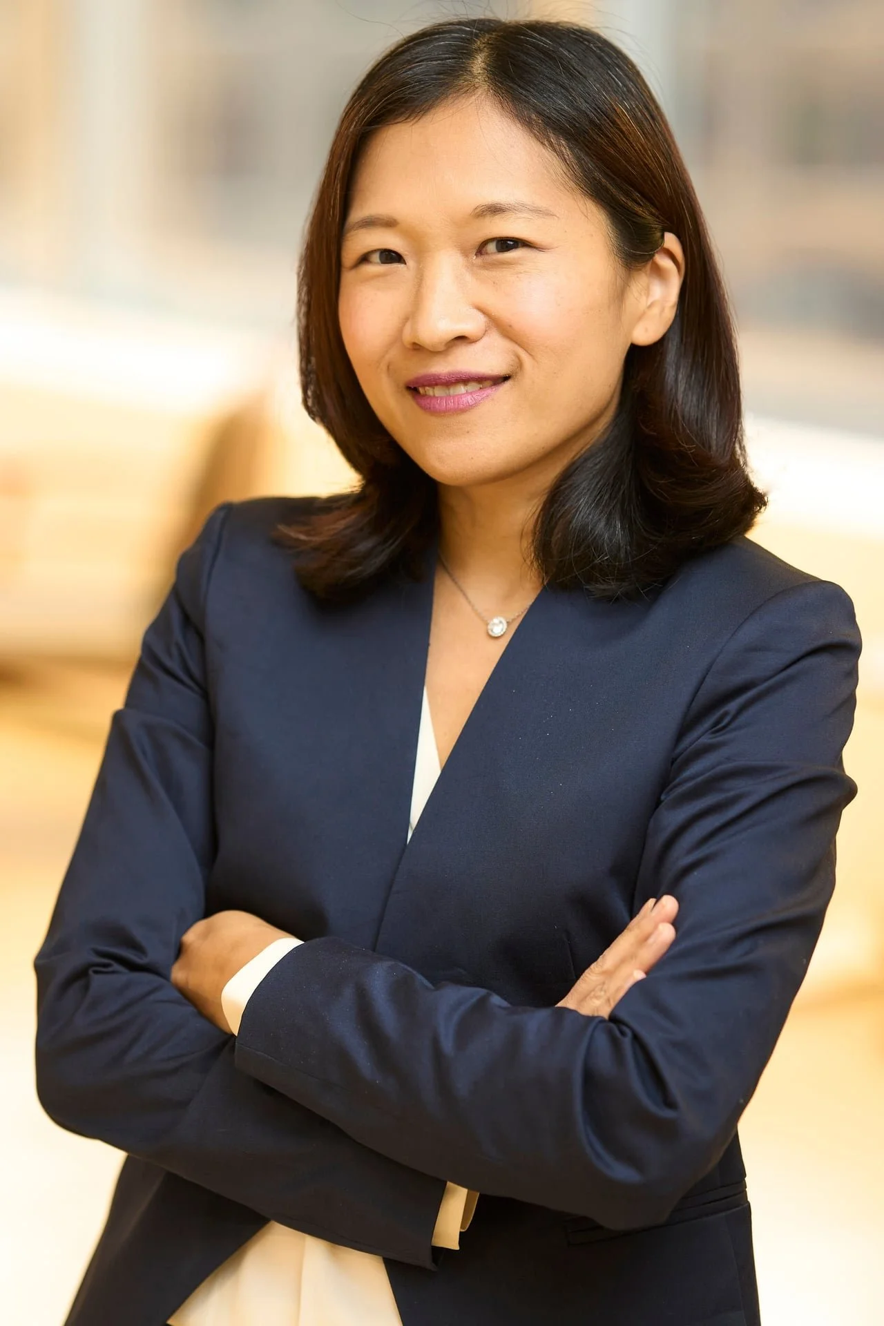 A professional woman with shoulder-length black hair wearing a dark blazer, smiling with crossed arms.