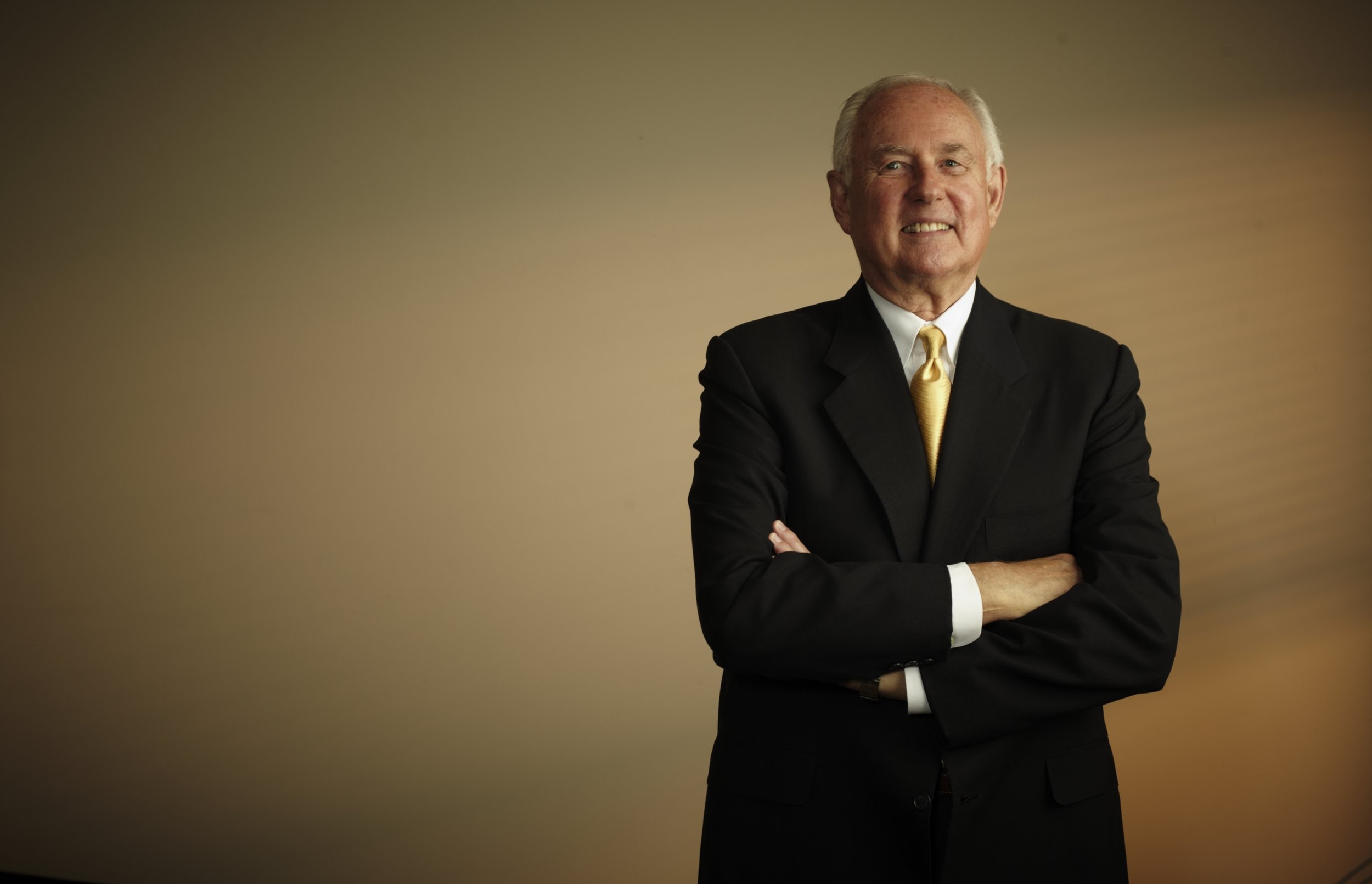 An older man with gray hair dressed in a black suit with a white shirt and a yellow tie, smiling and crossing his arms, standing against a plain beige background.