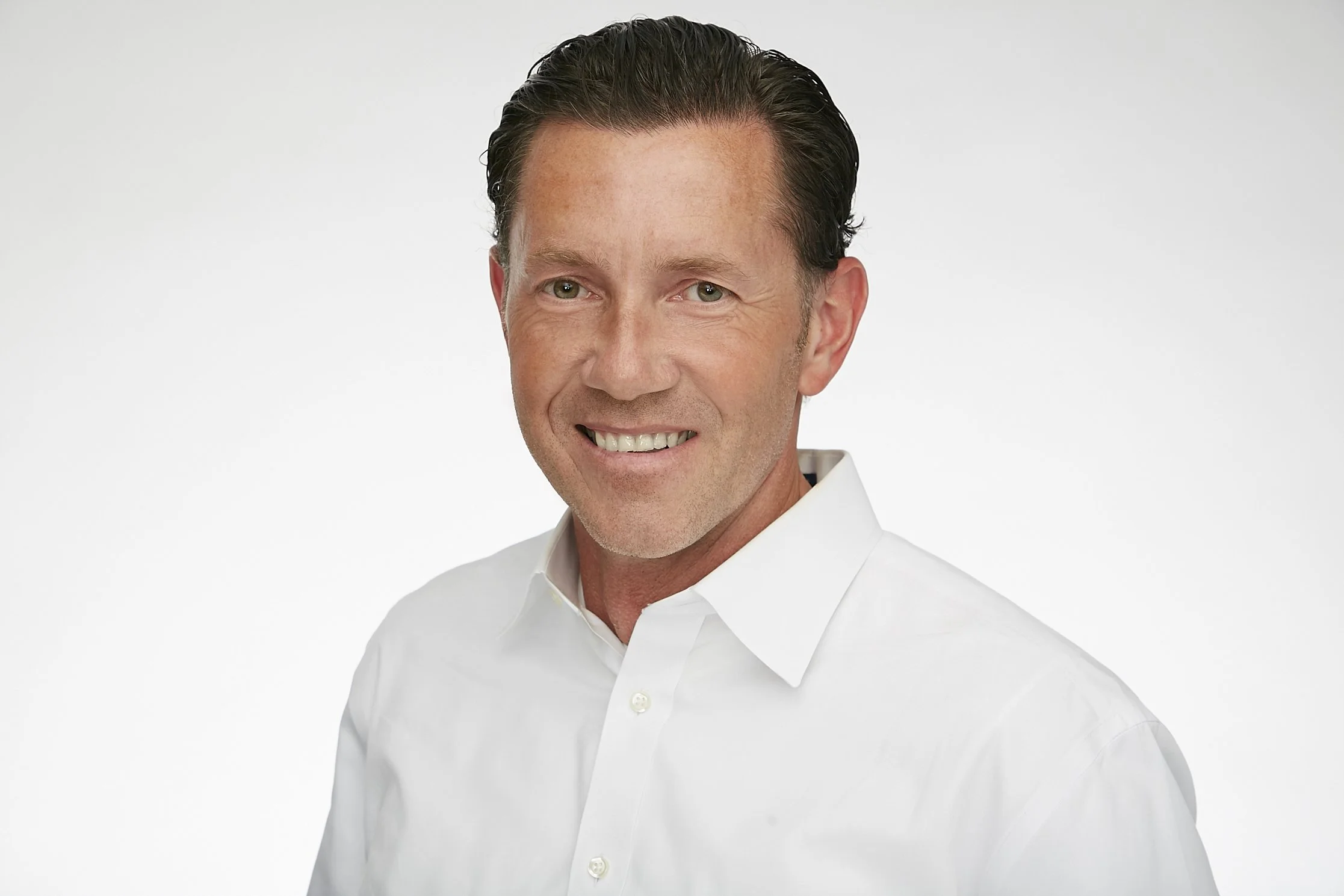 A smiling man with slicked-back dark hair, wearing a white dress shirt against a plain white background.