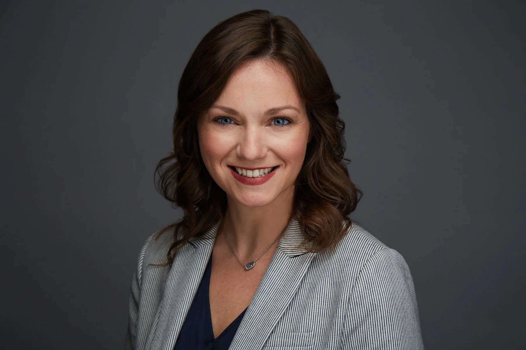 Portrait of a woman with shoulder-length wavy brown hair, blue eyes, wearing a grey striped blazer and a dark top, smiling against a dark grey background.