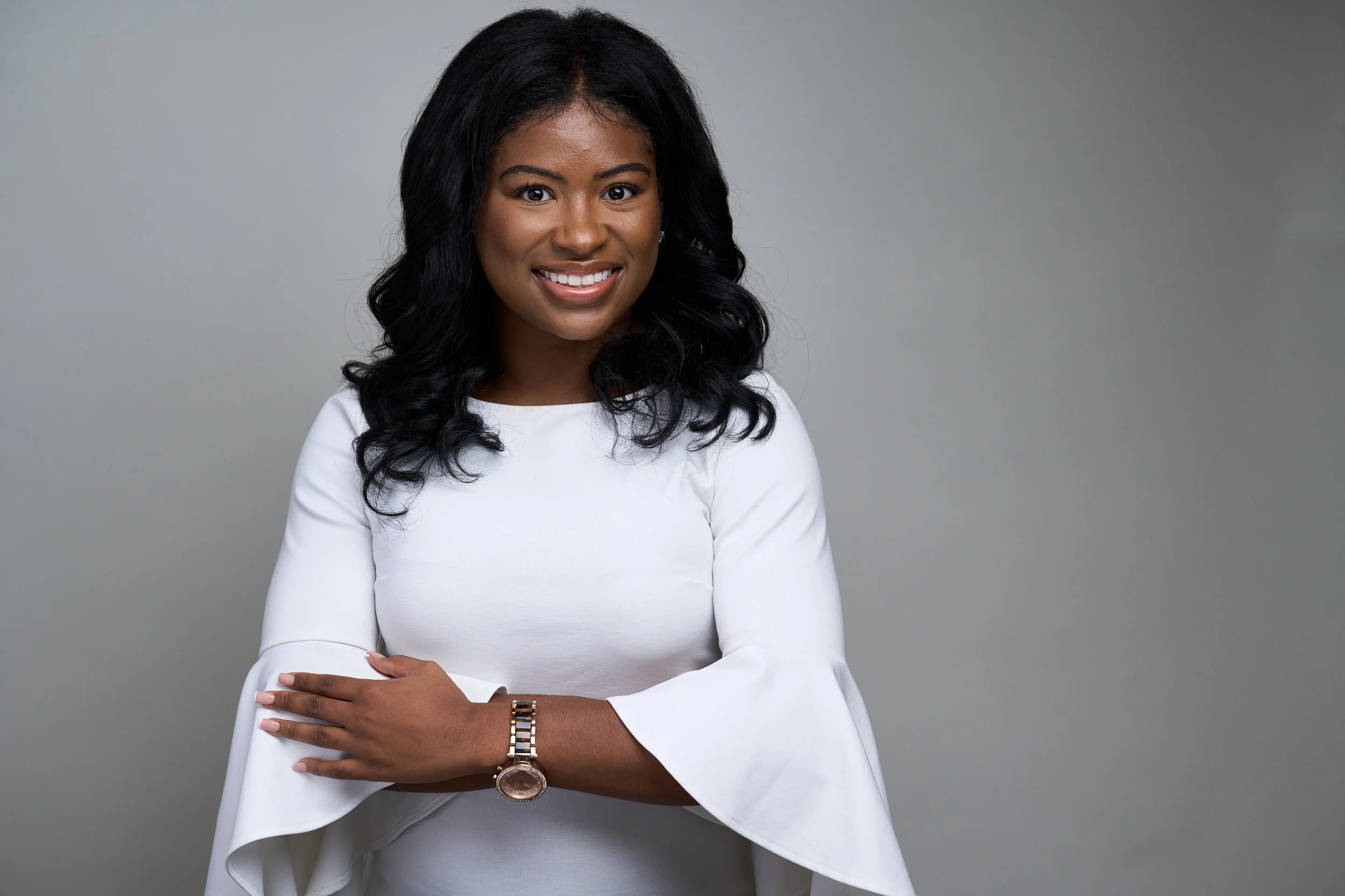 A woman with black, curly hair is smiling and looking at the camera. She is wearing a white top and has her arms crossed, with her left arm resting on her right arm. She is wearing a gold wristwatch and a gold bracelet on her left wrist. The backgrou