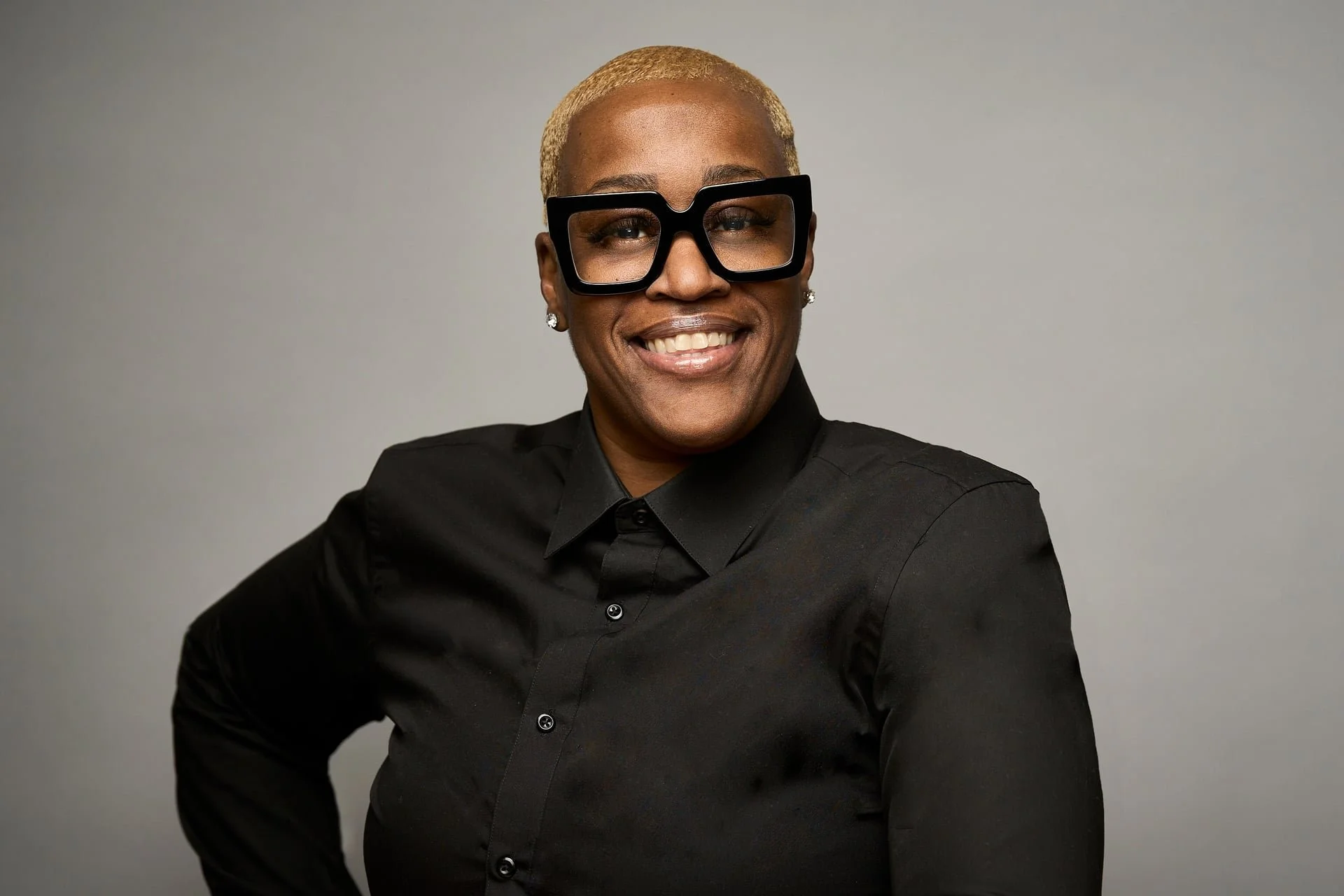 A smiling person with short blonde hair wearing large black glasses, glittery earrings, and a black button-up shirt against a plain gray background.