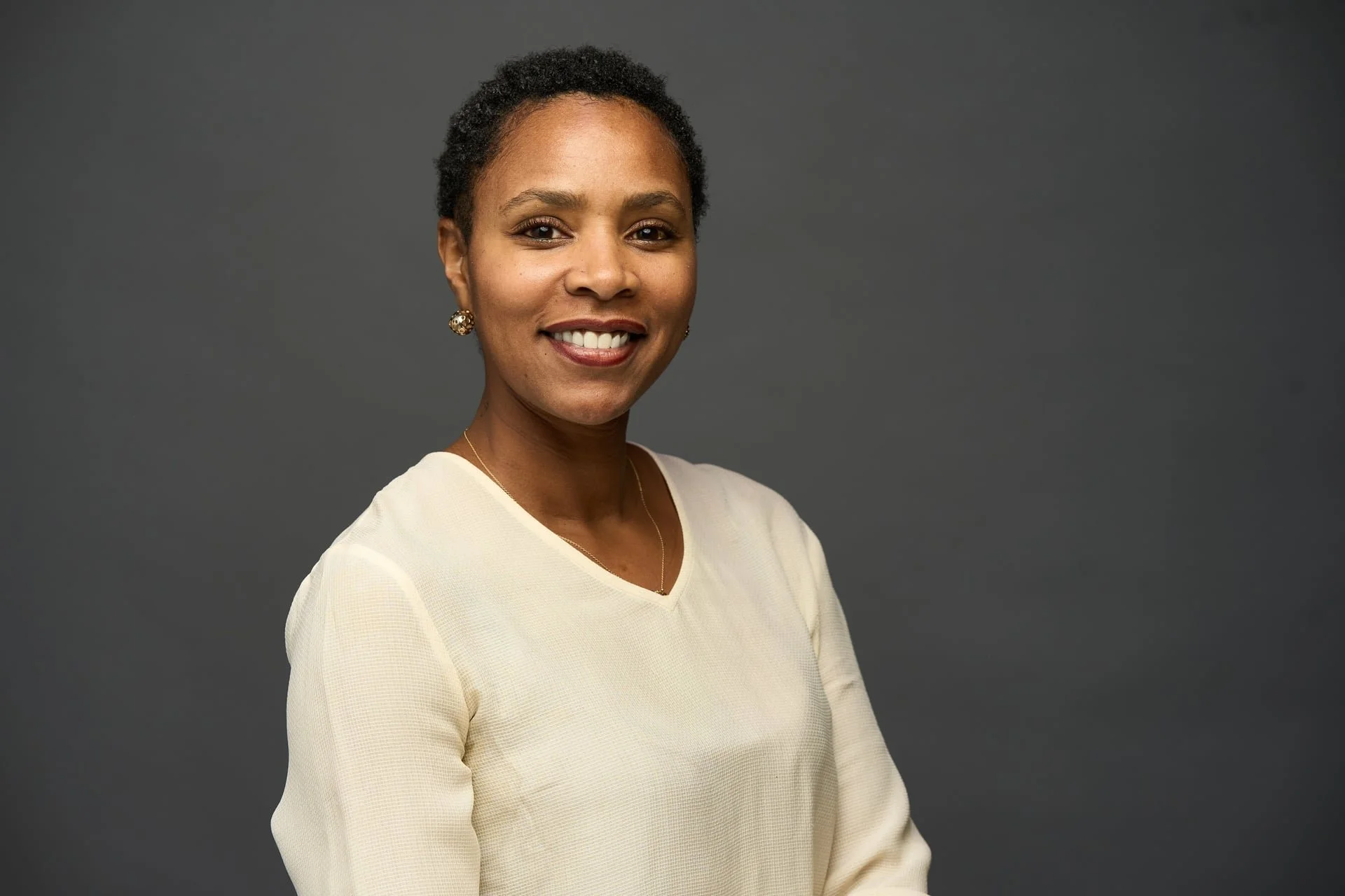 Portrait of a smiling Black woman with short, curly hair, wearing a cream-colored blouse and earrings, standing against a dark gray background.
