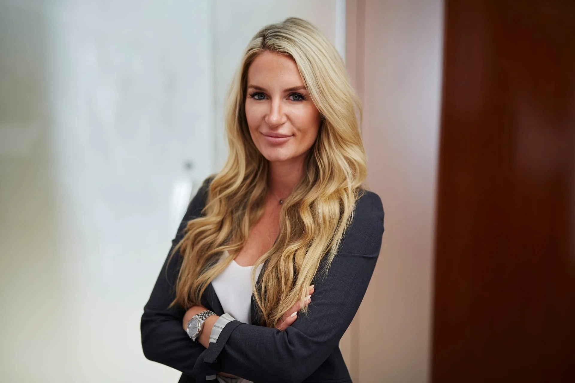 Confident woman in business attire with long blonde hair, crossed arms, standing indoors near a door.