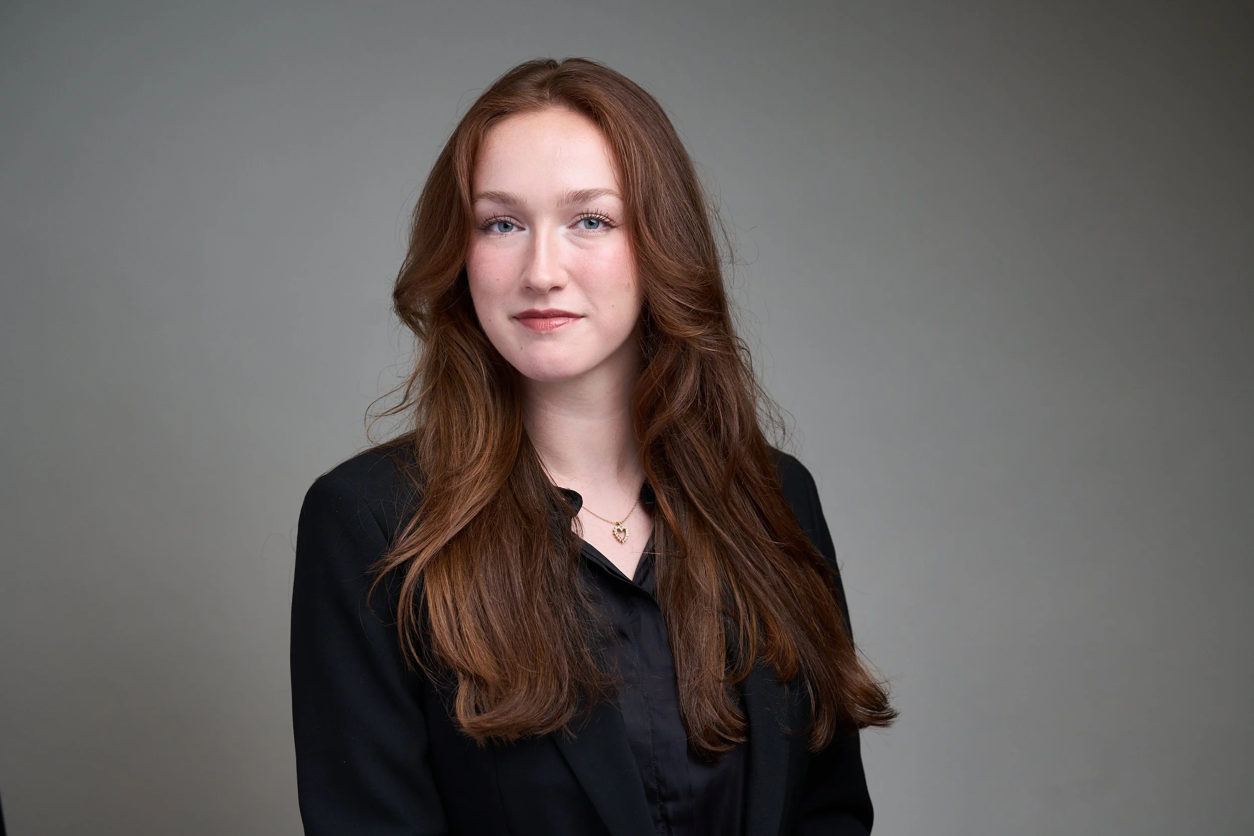 Headshot of a young woman with long wavy red hair, blue eyes, wearing a black top, a gold necklace with a heart pendant, against a plain gray background.