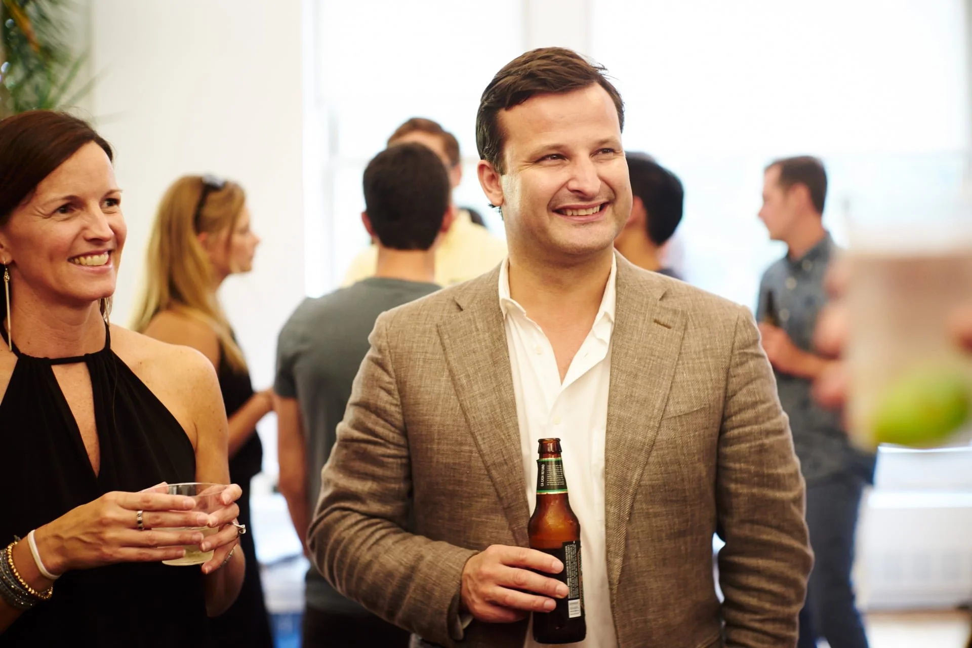 People socializing at a gathering, the man in the foreground is smiling and holding a beer bottle, while a woman next to him is smiling and holding a glass.