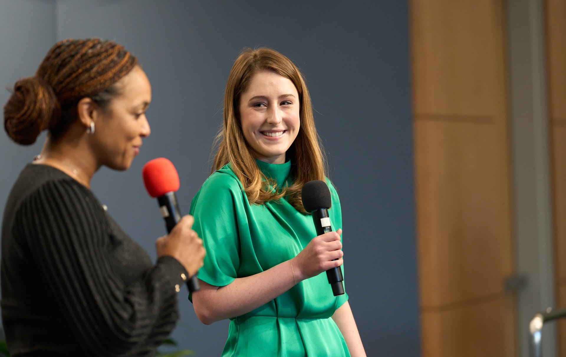 Two women are on stage holding microphones, one in a black top and the other in a green dress, engaging in a conversation with the woman in green smiling.
