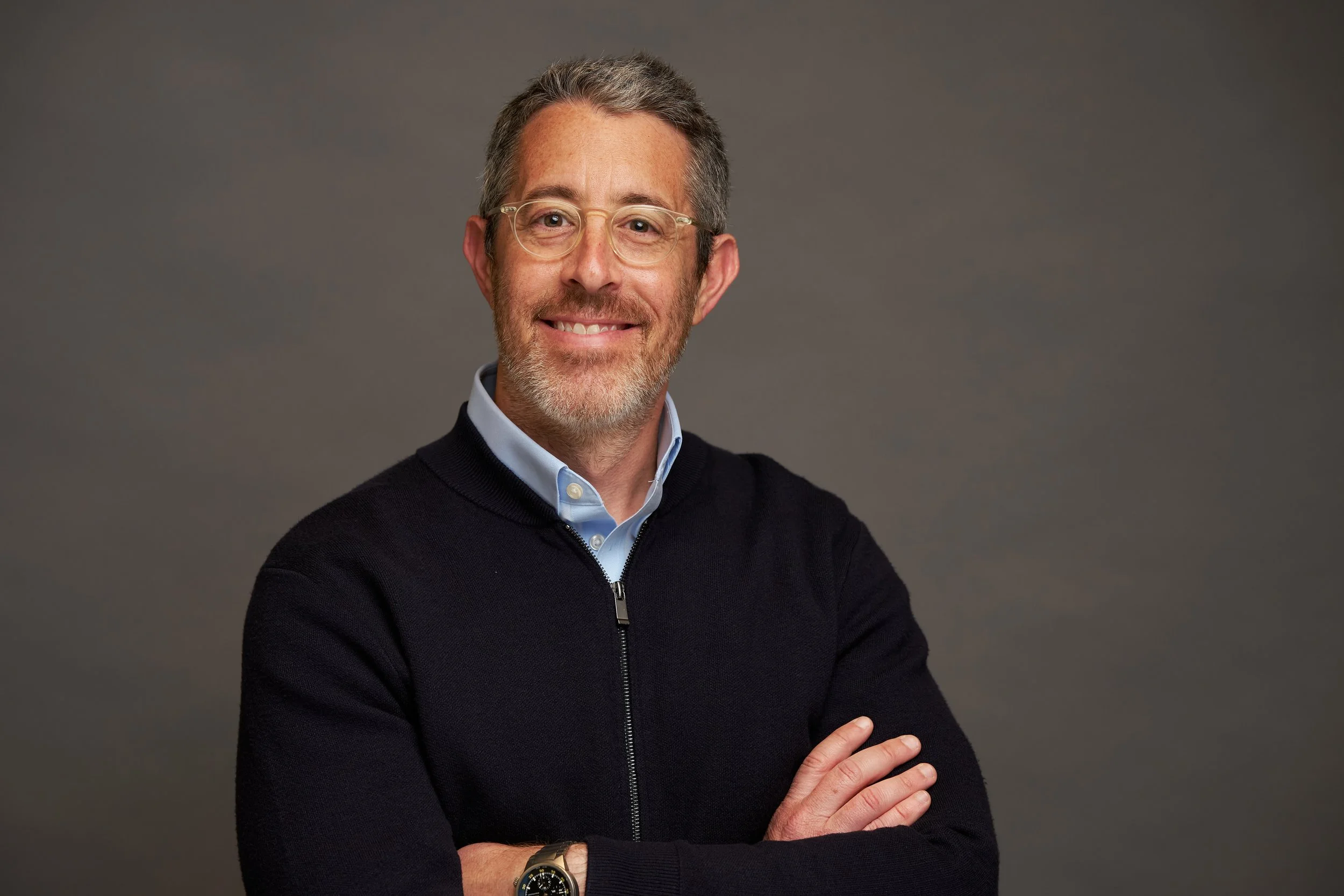 A middle-aged man smiling, wearing glasses, a light blue collared shirt, and a black sweater, with arms crossed against a plain gray background.