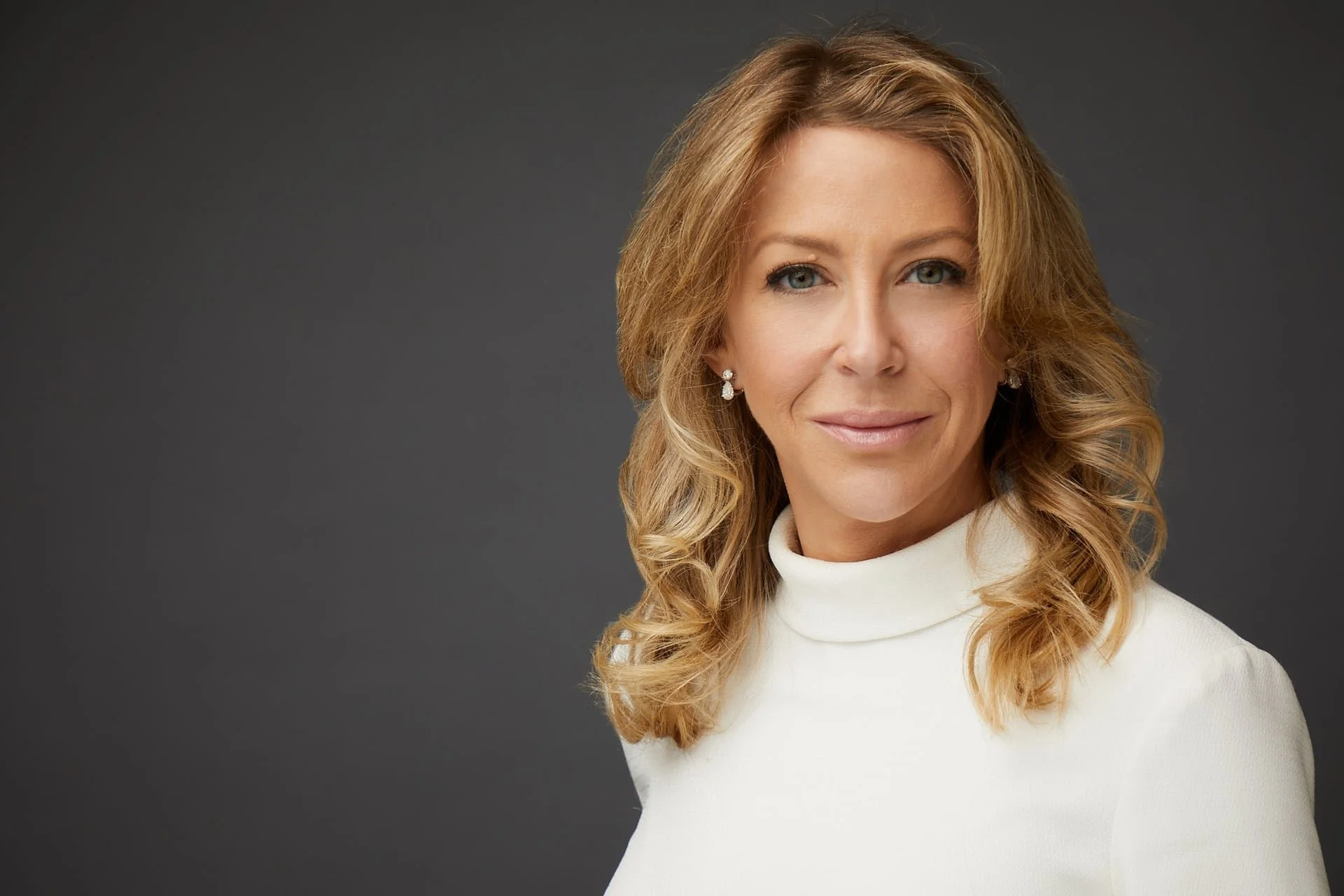 Portrait of a woman with blonde, wavy hair wearing pearl earrings and a white turtleneck sweater, smiling slightly, against a dark gray background.