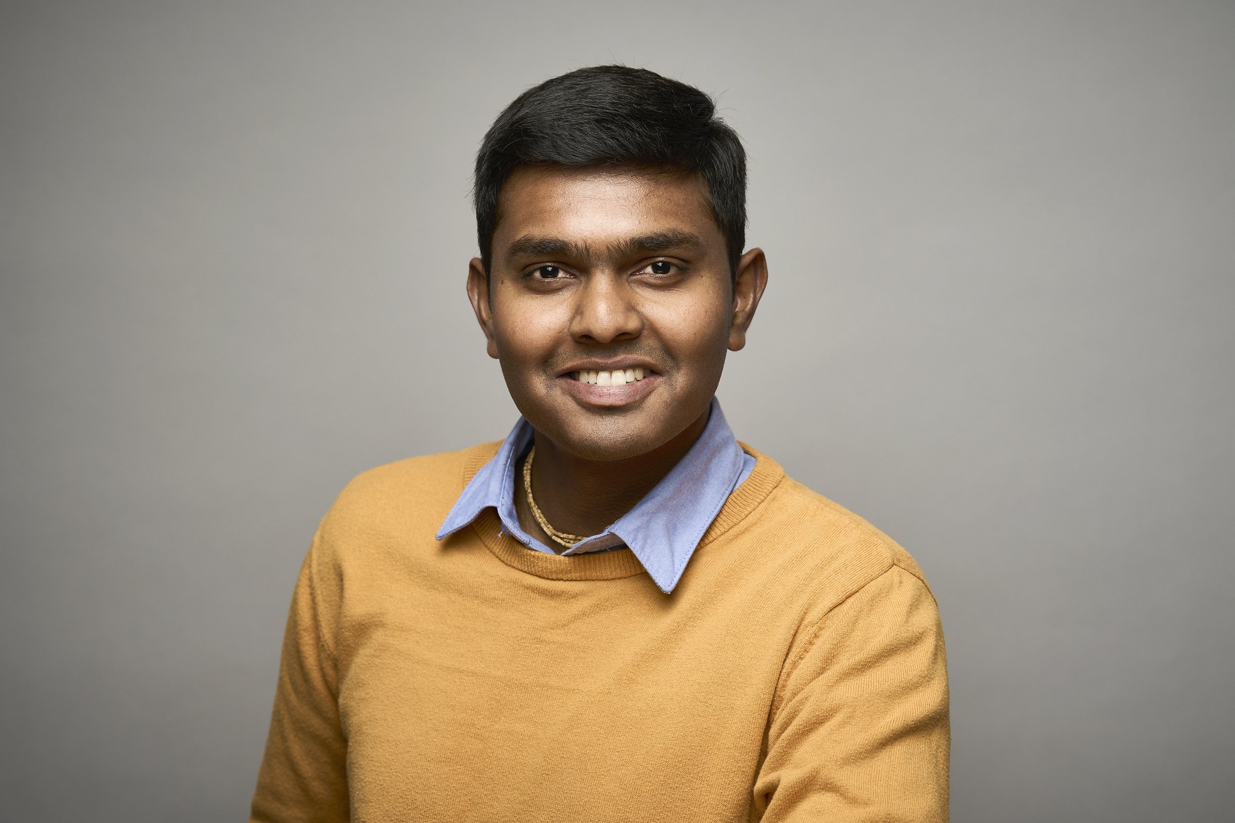 Portrait of a young man with dark hair, wearing a yellow sweater over a blue collared shirt, smiling lightly against a neutral gray background.