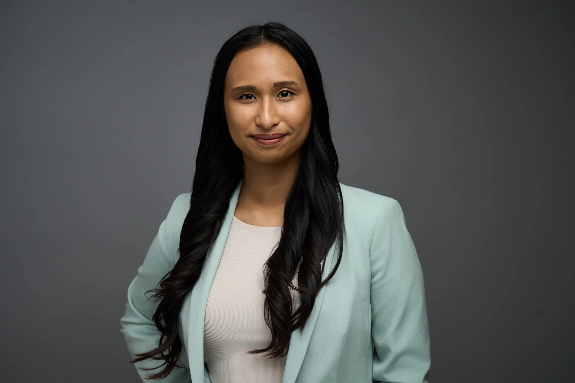 A young woman with long dark hair wearing a light green blazer and a white top, smiling slightly, standing against a dark gray background.