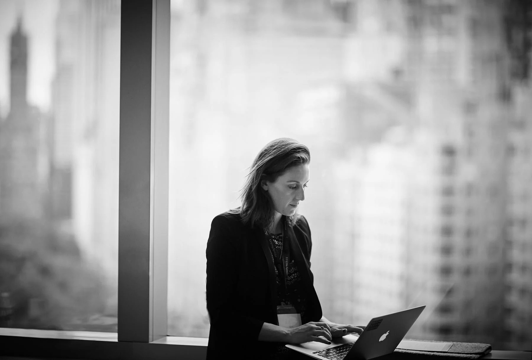 A woman with shoulder-length hair working on a laptop by a large window in black and white.