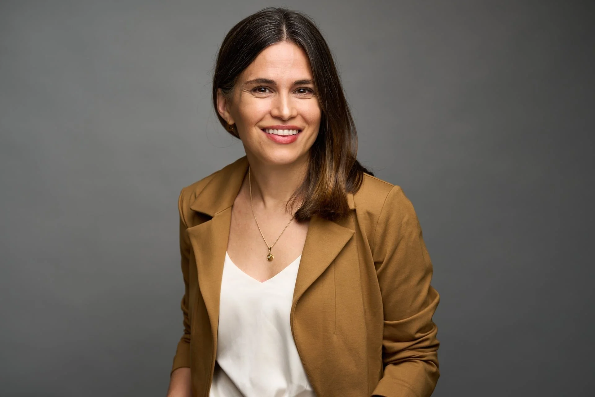 Portrait of a woman with shoulder-length brown hair, wearing a tan blazer over a white top, smiling against a gray background.