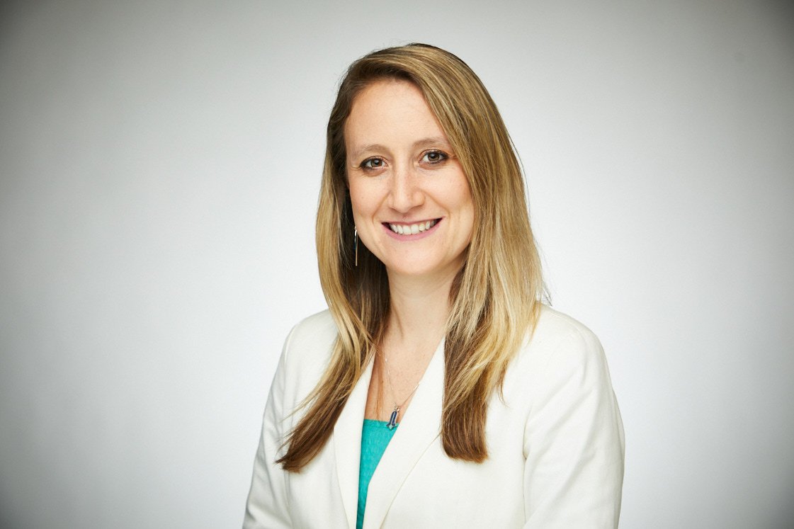 A woman with long, light brown hair wearing a white blazer and turquoise top, smiling against a plain background.