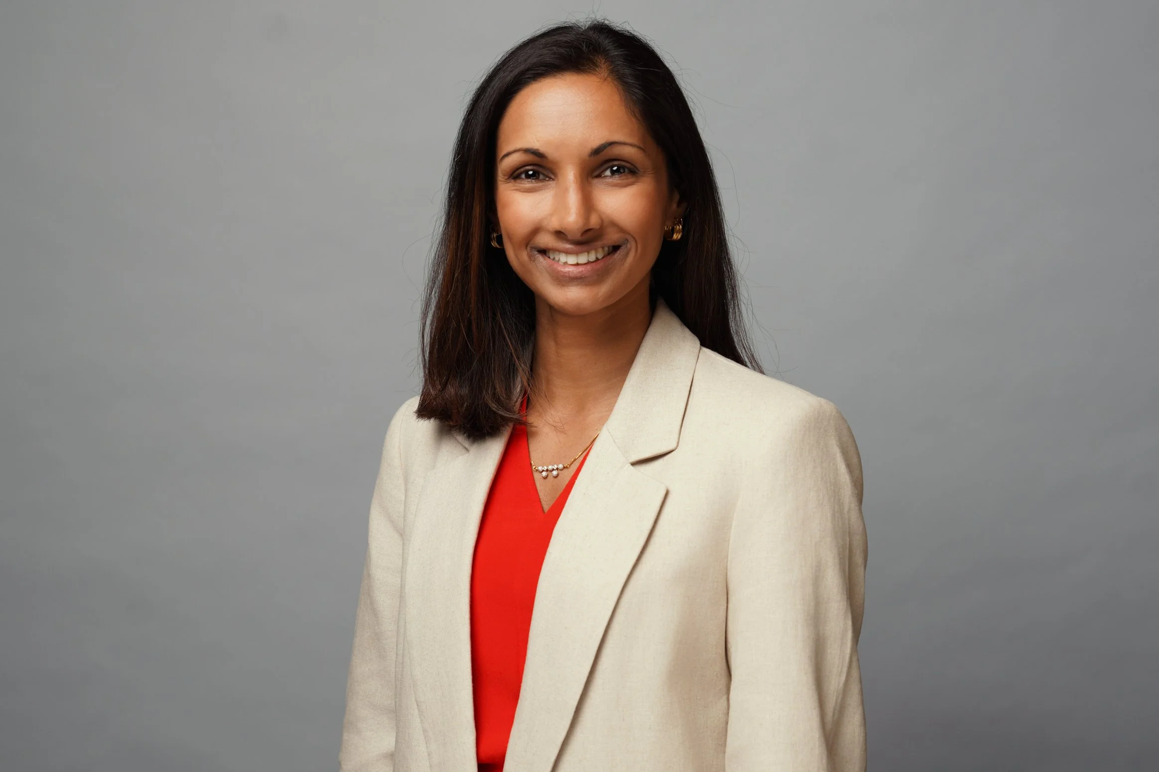 Portrait of a woman with long dark hair wearing a beige blazer over a red top, smiling in front of a plain gray background.