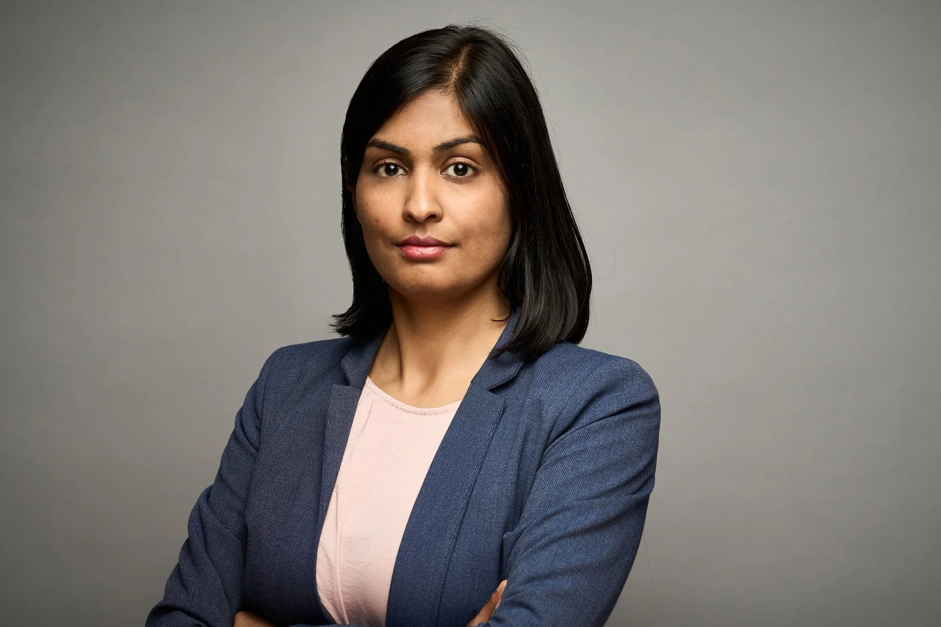 A professional woman with shoulder-length black hair, wearing a navy blazer and light pink shirt, standing with her arms crossed against a plain gray background.