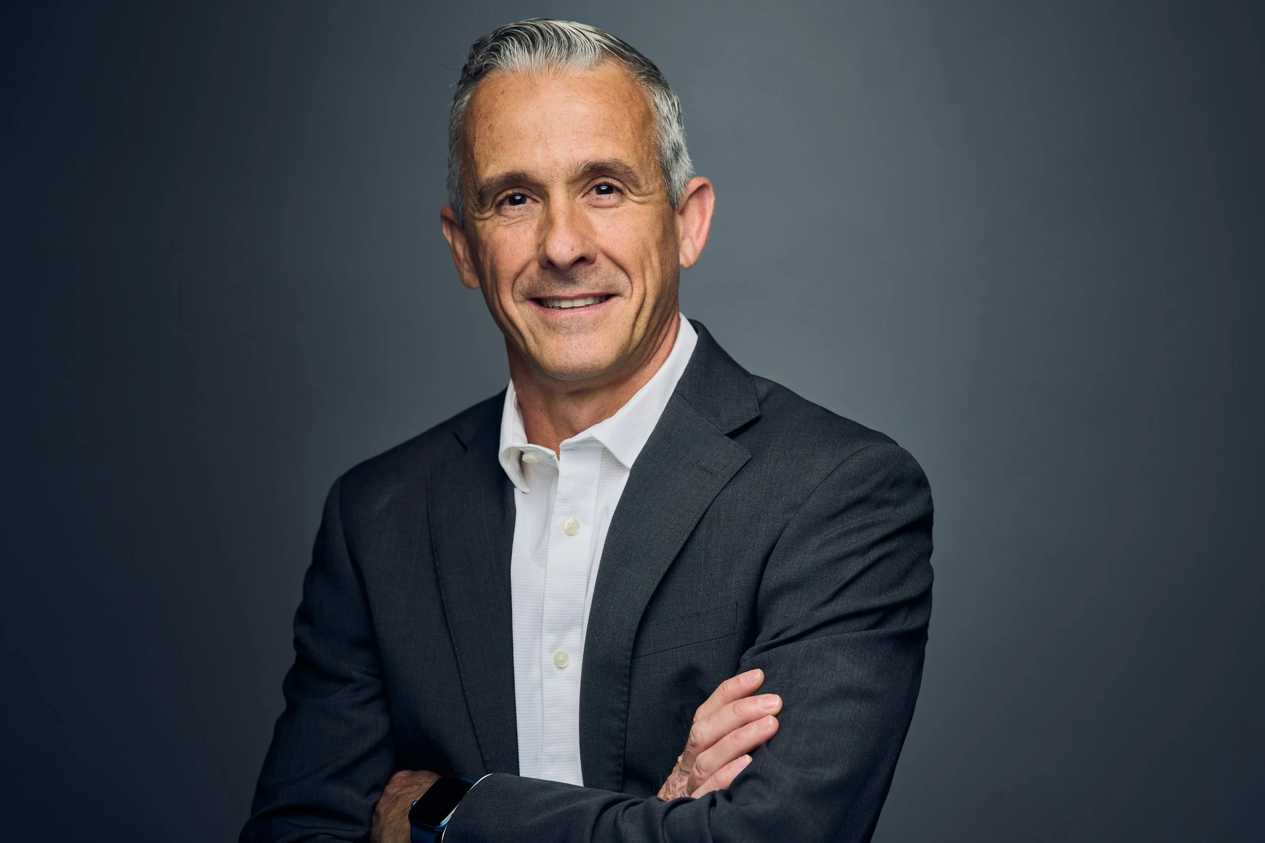 A middle-aged man with gray hair wearing a dark suit jacket, white shirt, and a black wristband, smiling and crossing his arms against a gray background.