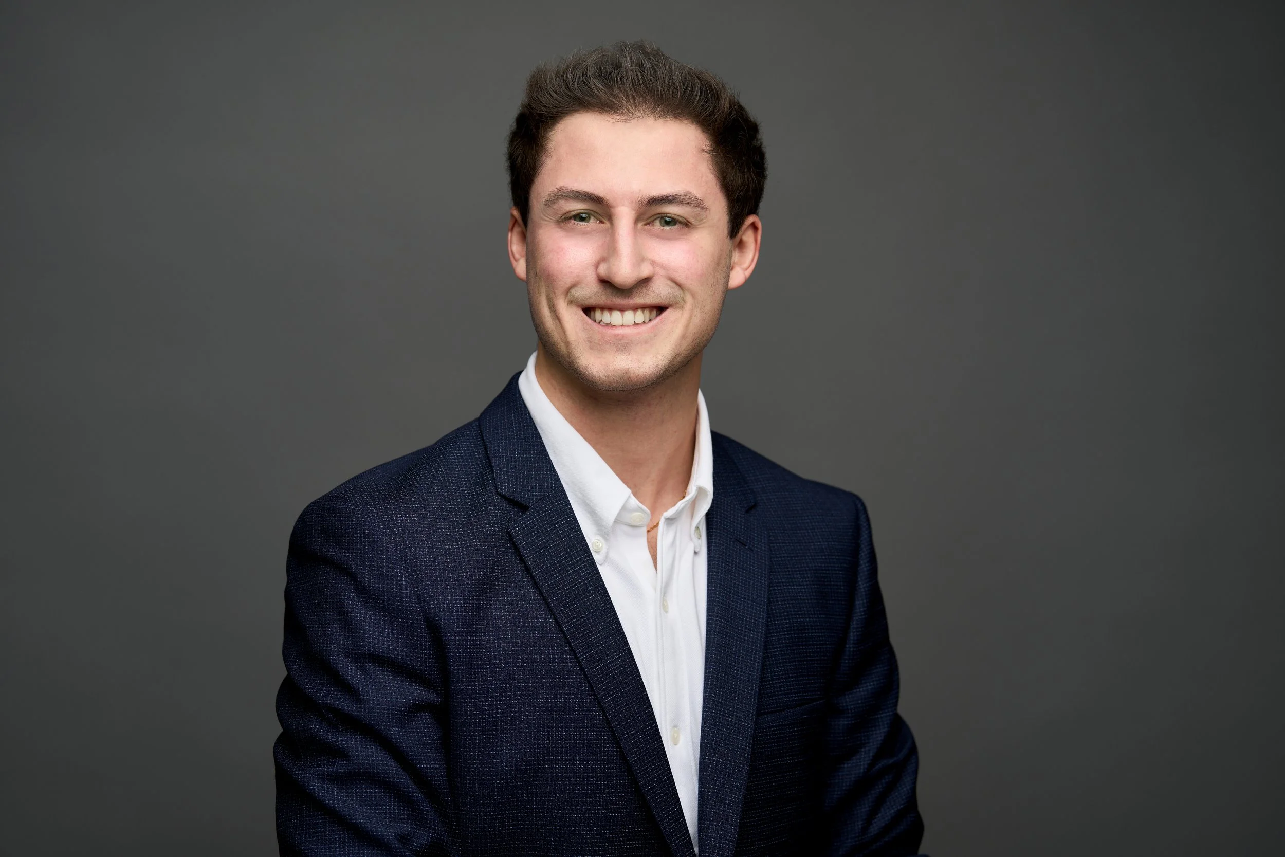 A young man with dark hair, wearing a dark blue suit jacket and white shirt, smiling against a plain gray background.