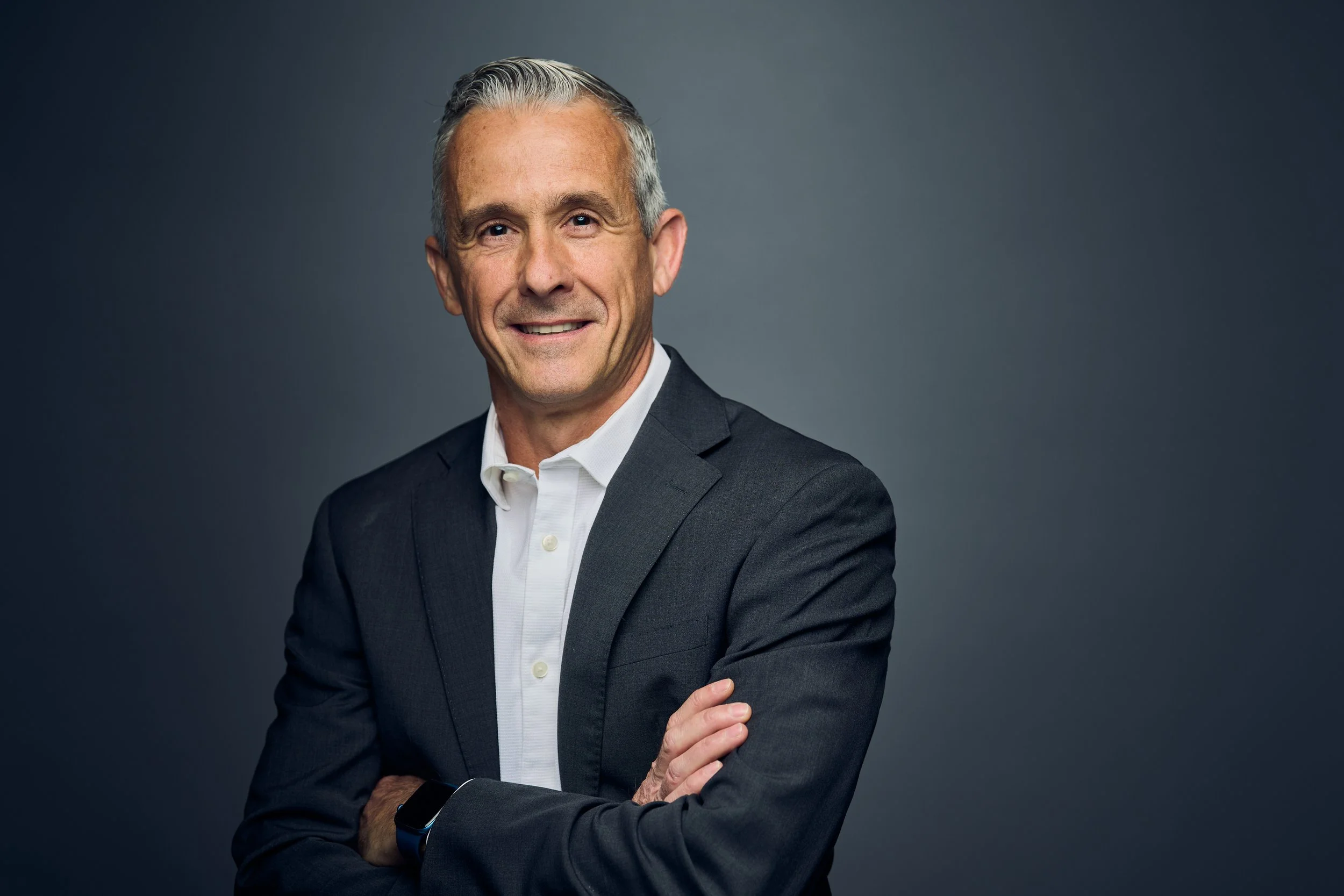 A mature man with gray hair, wearing a dark suit and white shirt, standing against a gray background with arms crossed and smiling.
