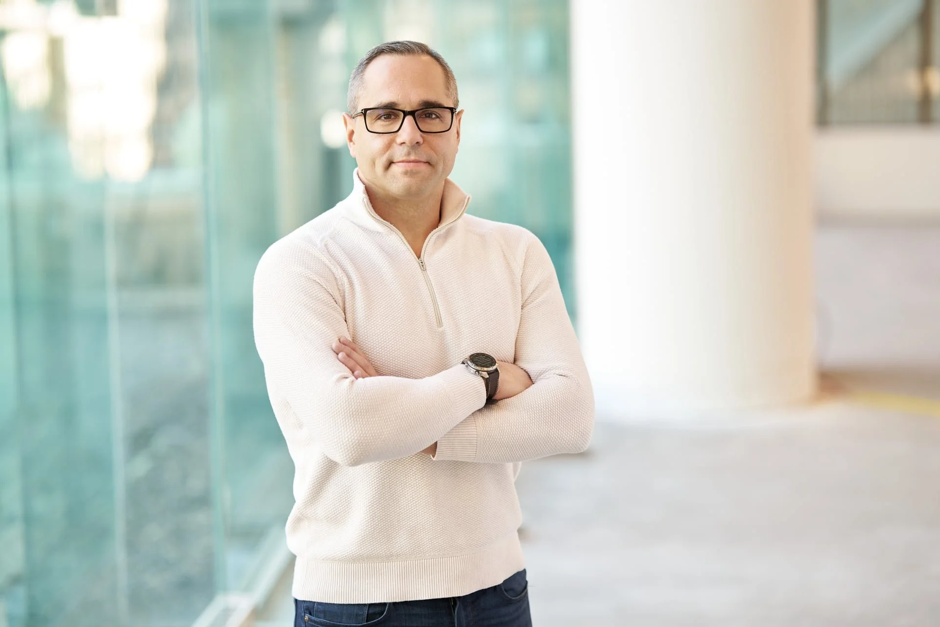 A man wearing glasses and a beige sweater with arms crossed, standing indoors near a glass window with a blurred background.