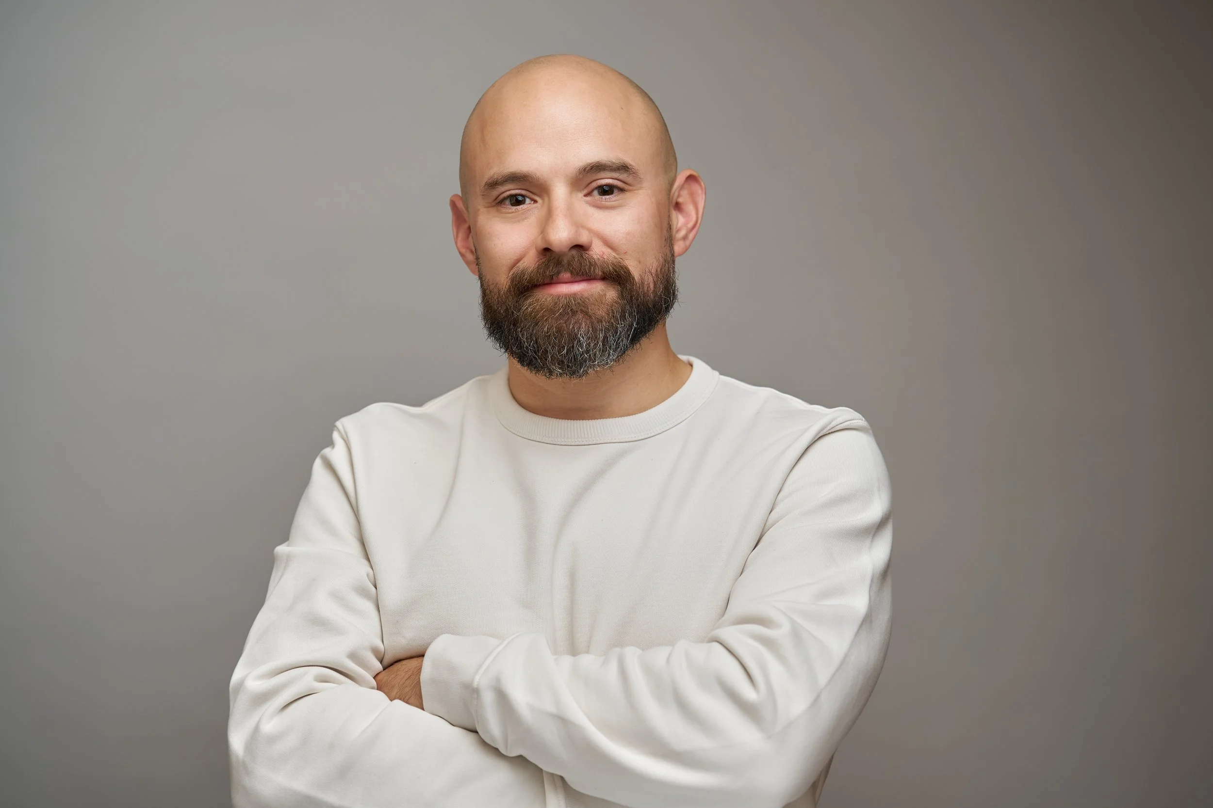 A man with a bald head, full beard, and mustache wearing a white long-sleeve shirt, crossing his arms and looking at the camera against a gray background.