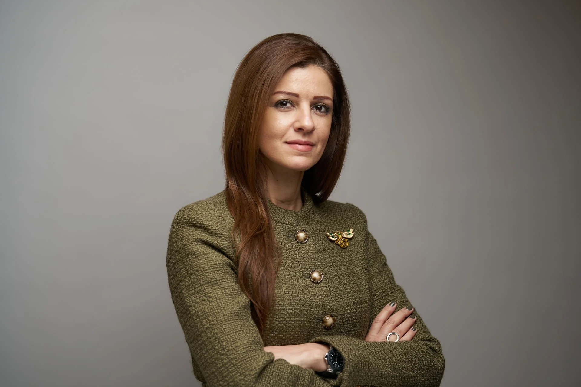 A woman with long brown hair wearing a green textured blazer with gold and animal-shaped brooches, arms crossed, against a plain gray background.