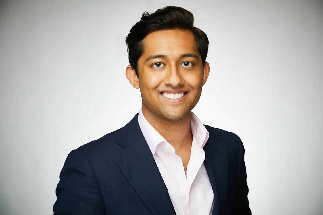 Professional portrait of a smiling man in a dark suit and white shirt against a plain light background.