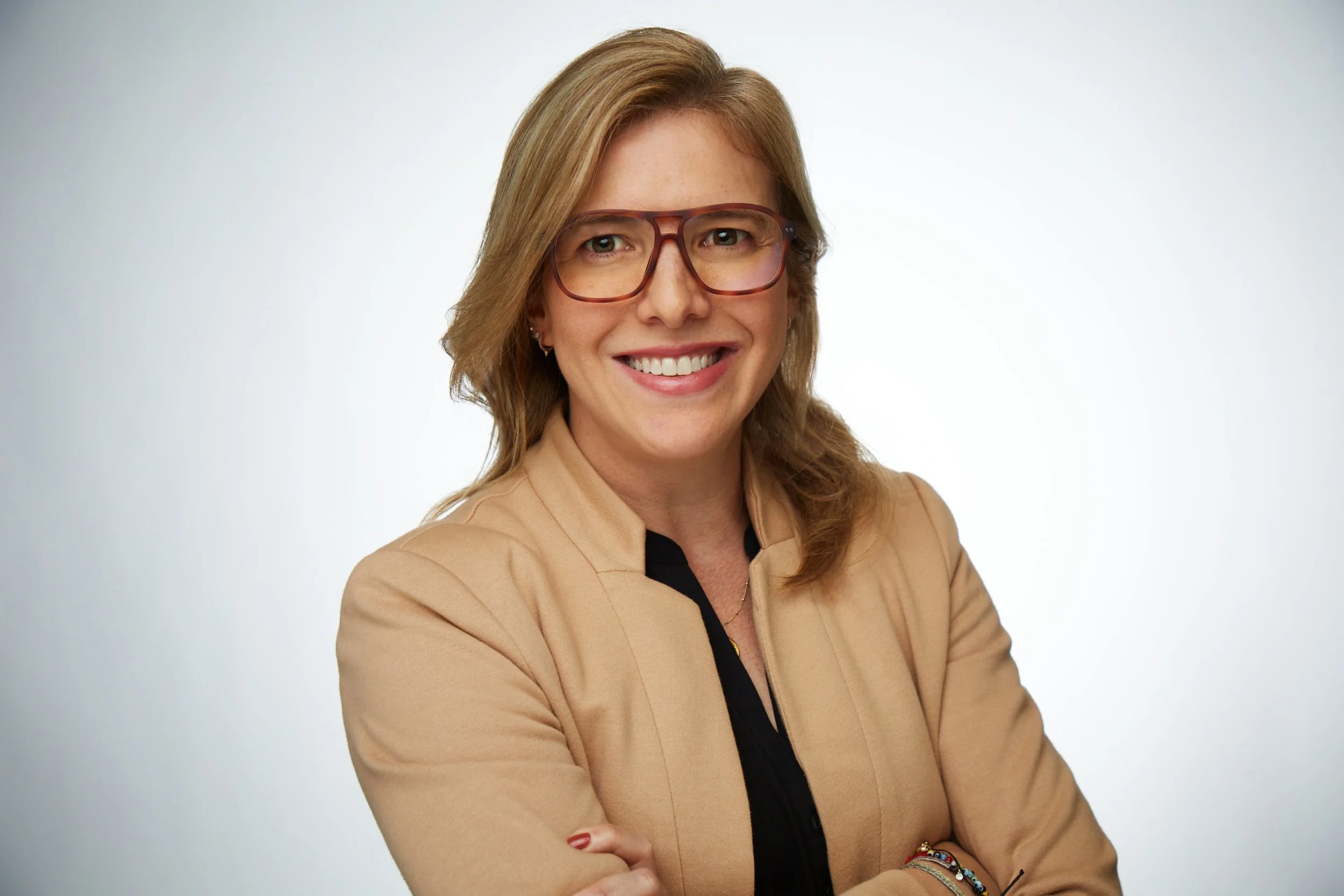 A woman with shoulder-length light brown hair wearing large brown glasses, a beige blazer, and a black top, smiling and crossing her arms, against a plain white background.