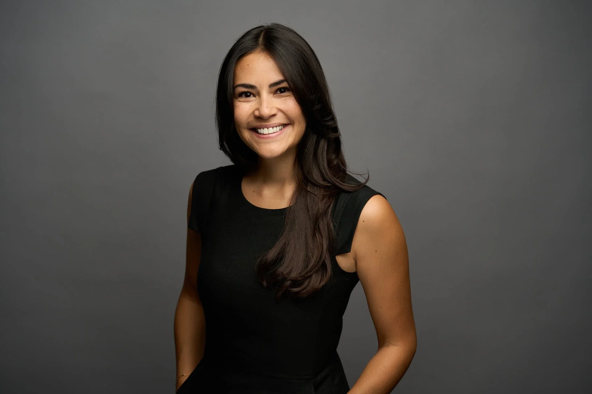 Portrait of a smiling woman with long dark hair, wearing a black sleeveless top, standing against a plain gray background.