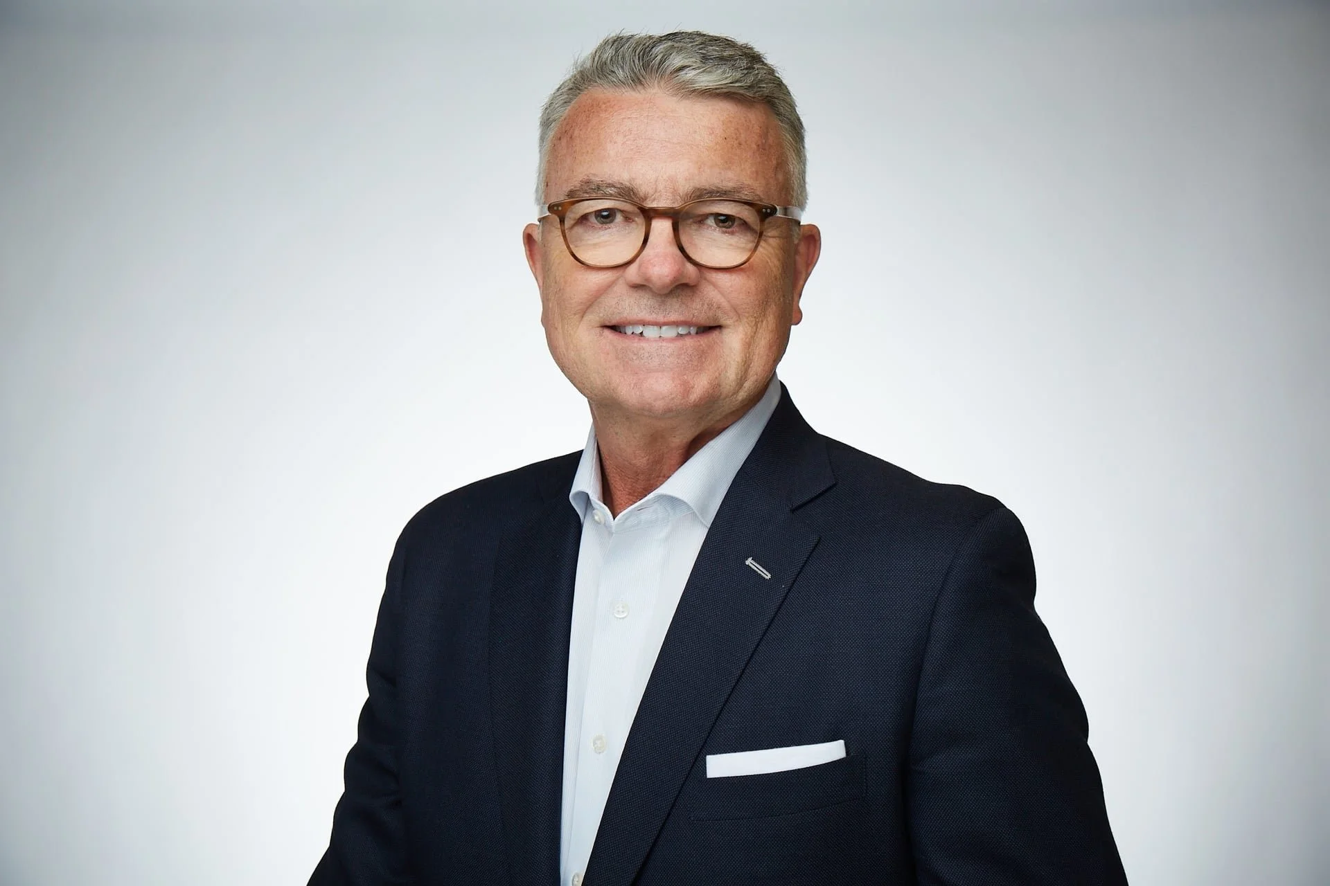 A middle-aged man with gray hair and glasses, wearing a dark suit and white shirt, smiling at the camera against a plain light background.