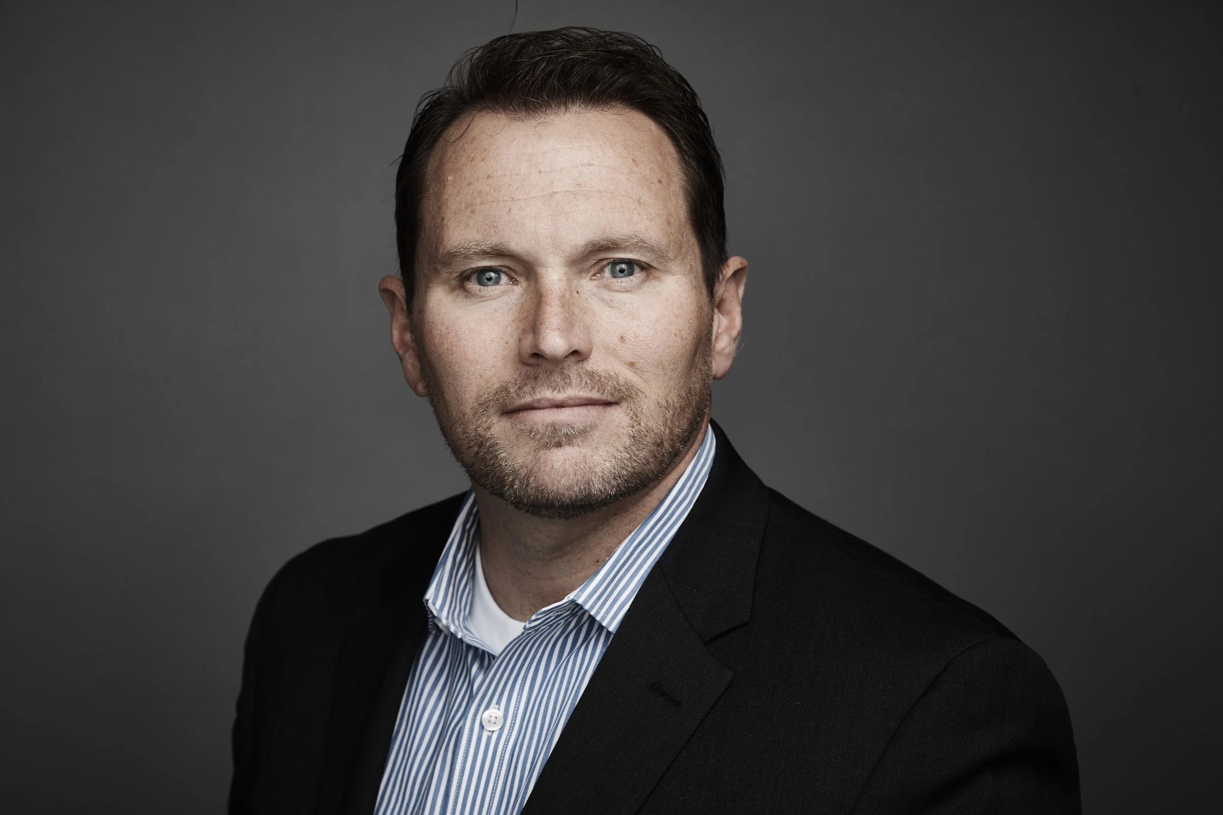 A professional headshot of a man with blue eyes and brown hair, wearing a black blazer and a blue and white striped shirt, against a gray background.