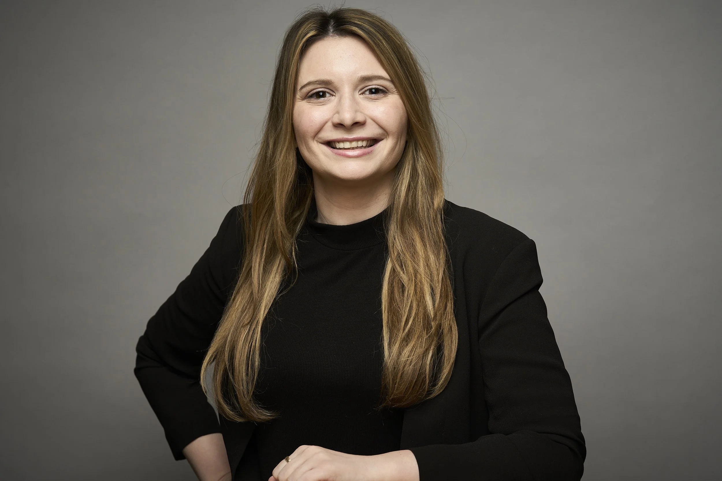A woman with long, light brown hair wearing a black turtleneck, smiling against a plain gray background.