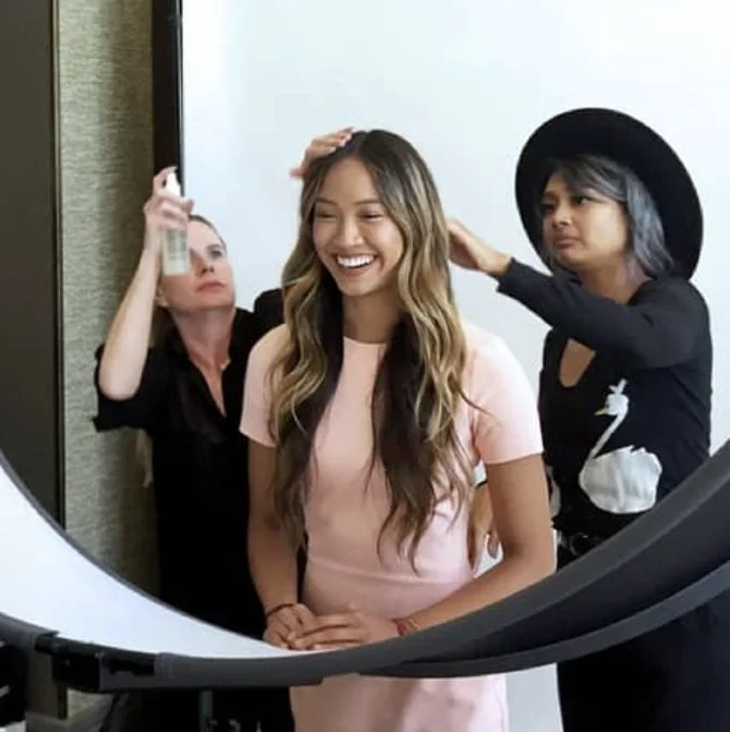 A young woman in a pink dress smiles as two stylists, one with dark hair and a black hat and the other with a black shirt, do her hair and makeup in front of a white backdrop.