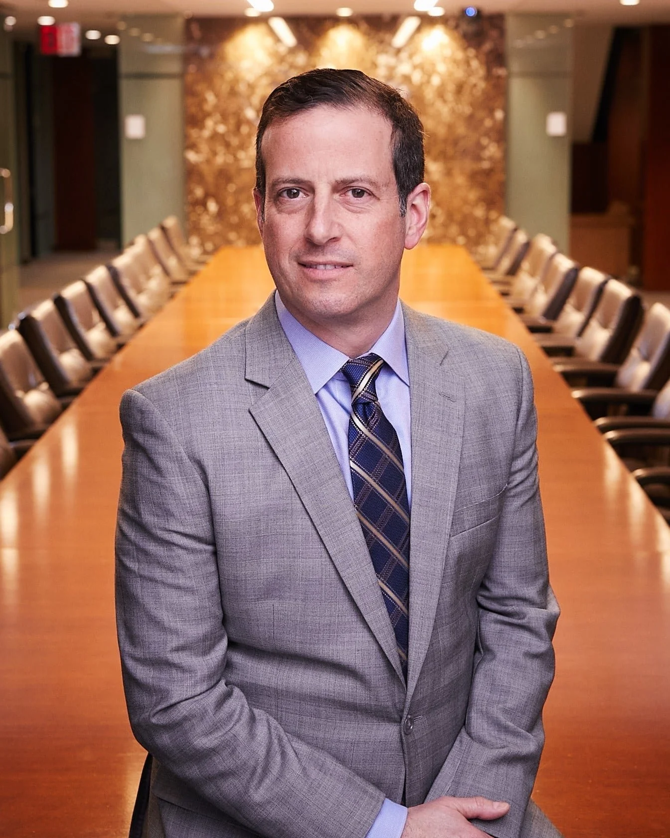 A man wearing a gray suit with a blue dress shirt and a striped tie, standing in a conference room with a long table and chairs, with a textured wall in the background.
