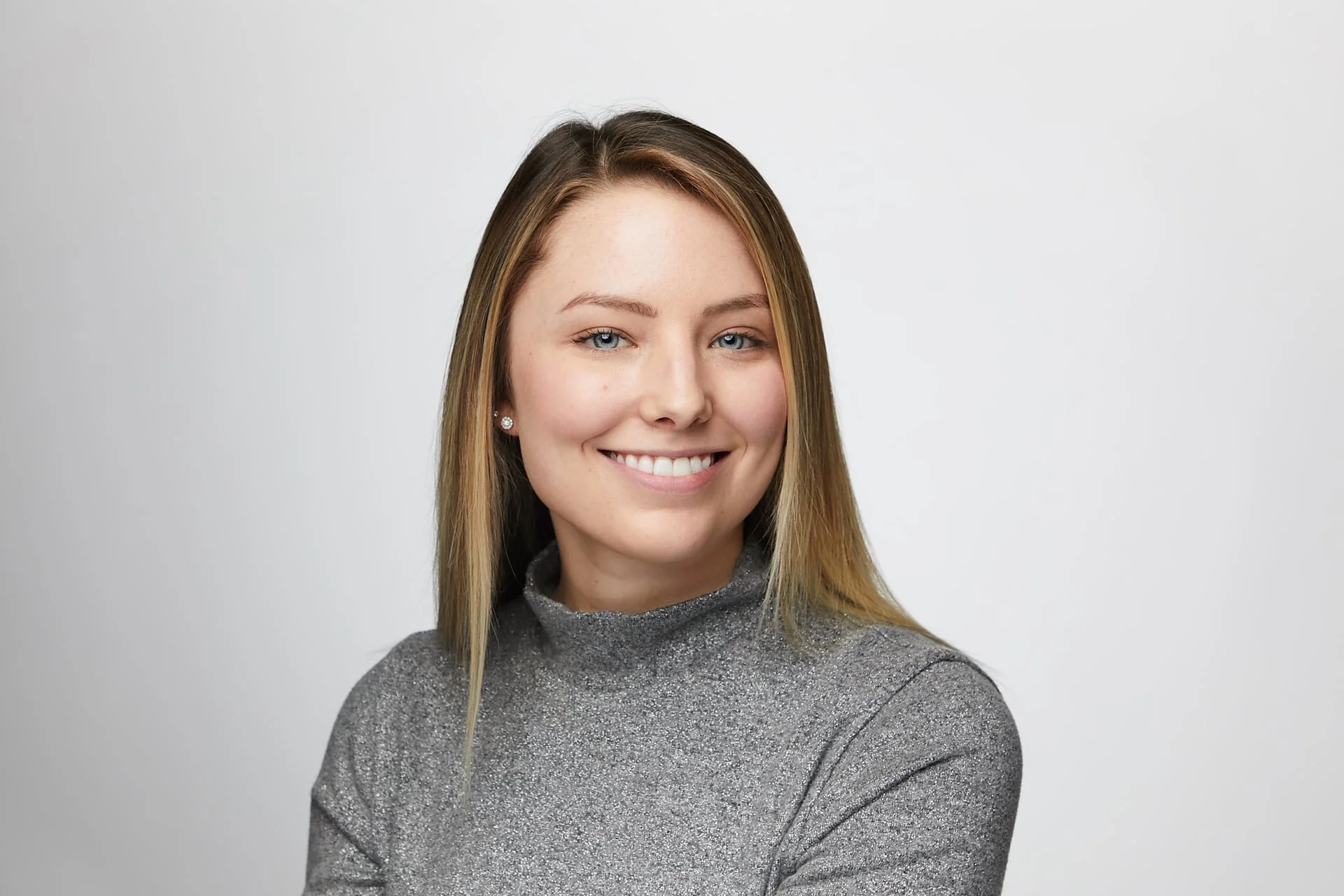 A young woman with blonde hair and blue eyes, smiling, wearing a gray, glittery turtleneck sweater, posed against a plain white background.