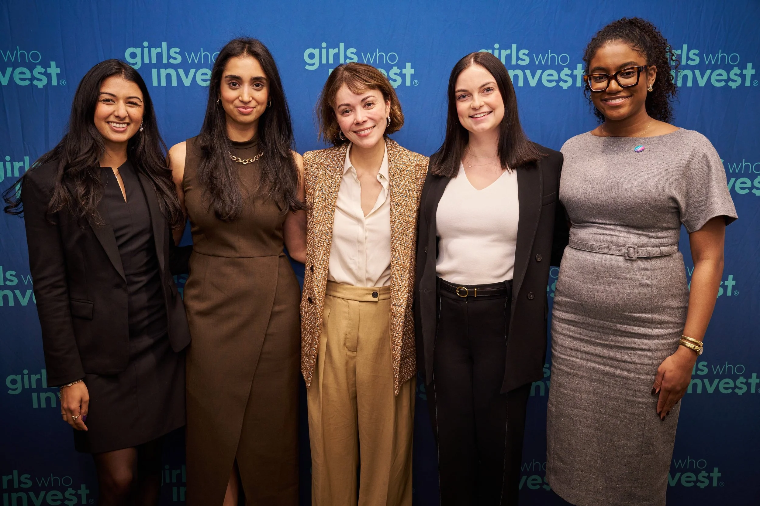 Group of five women standing together at an event with a blue backdrop that reads 'girls who invest' multiple times.