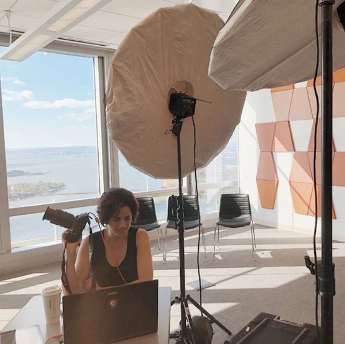 A woman sitting at a desk in a bright office with large windows overlooking a body of water and a cityscape, holding a camera, with professional lighting equipment setup for a photo or video shoot.