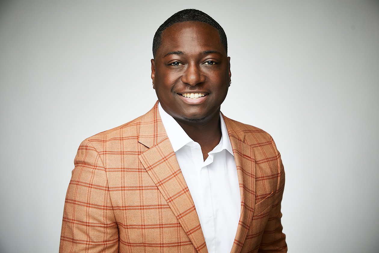 Professional portrait of a smiling Black man in a brown plaid suit jacket and white shirt against a plain light gray background.