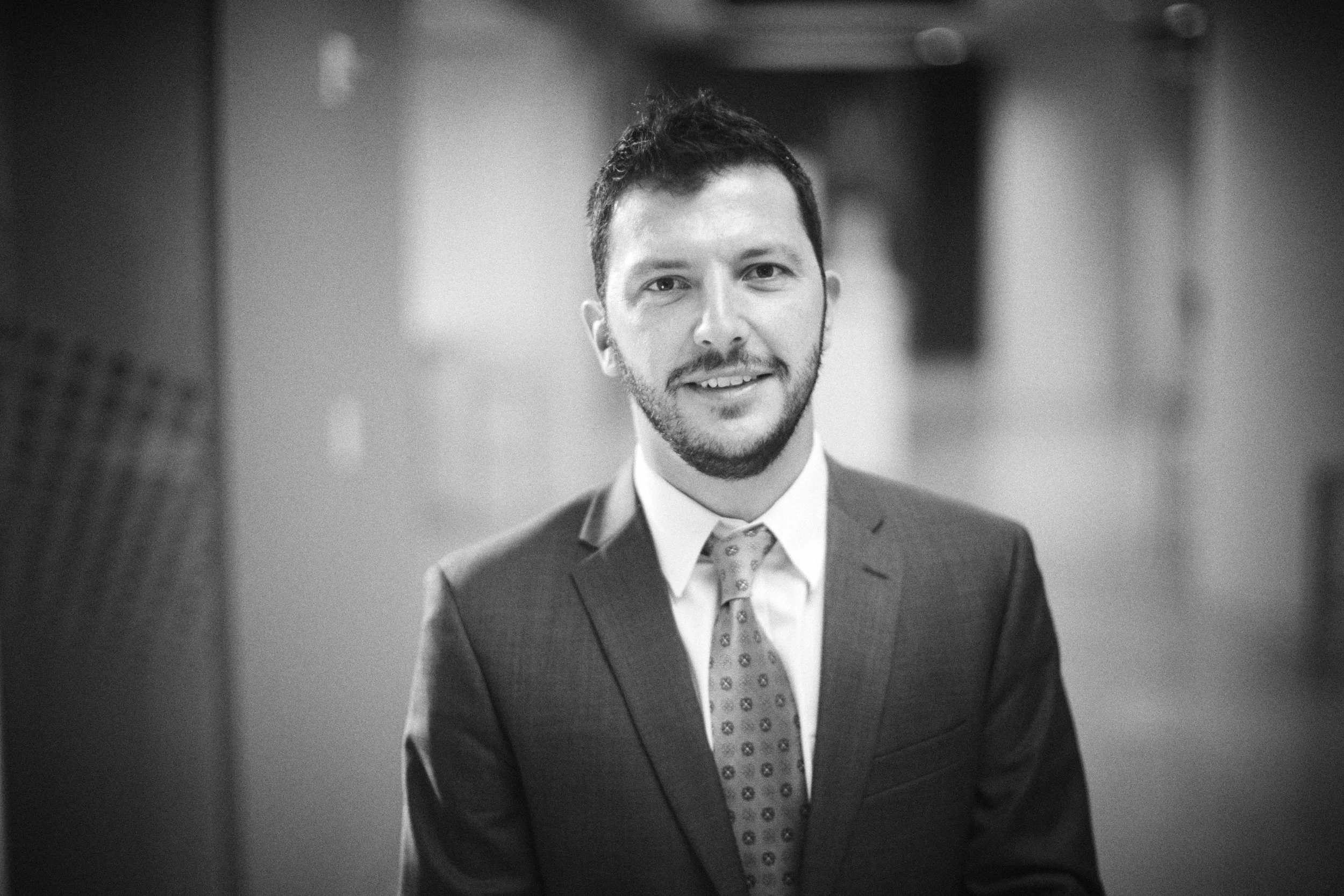 A black and white portrait of a young man wearing a suit, tie, and dress shirt, smiling slightly. The background is blurred.
